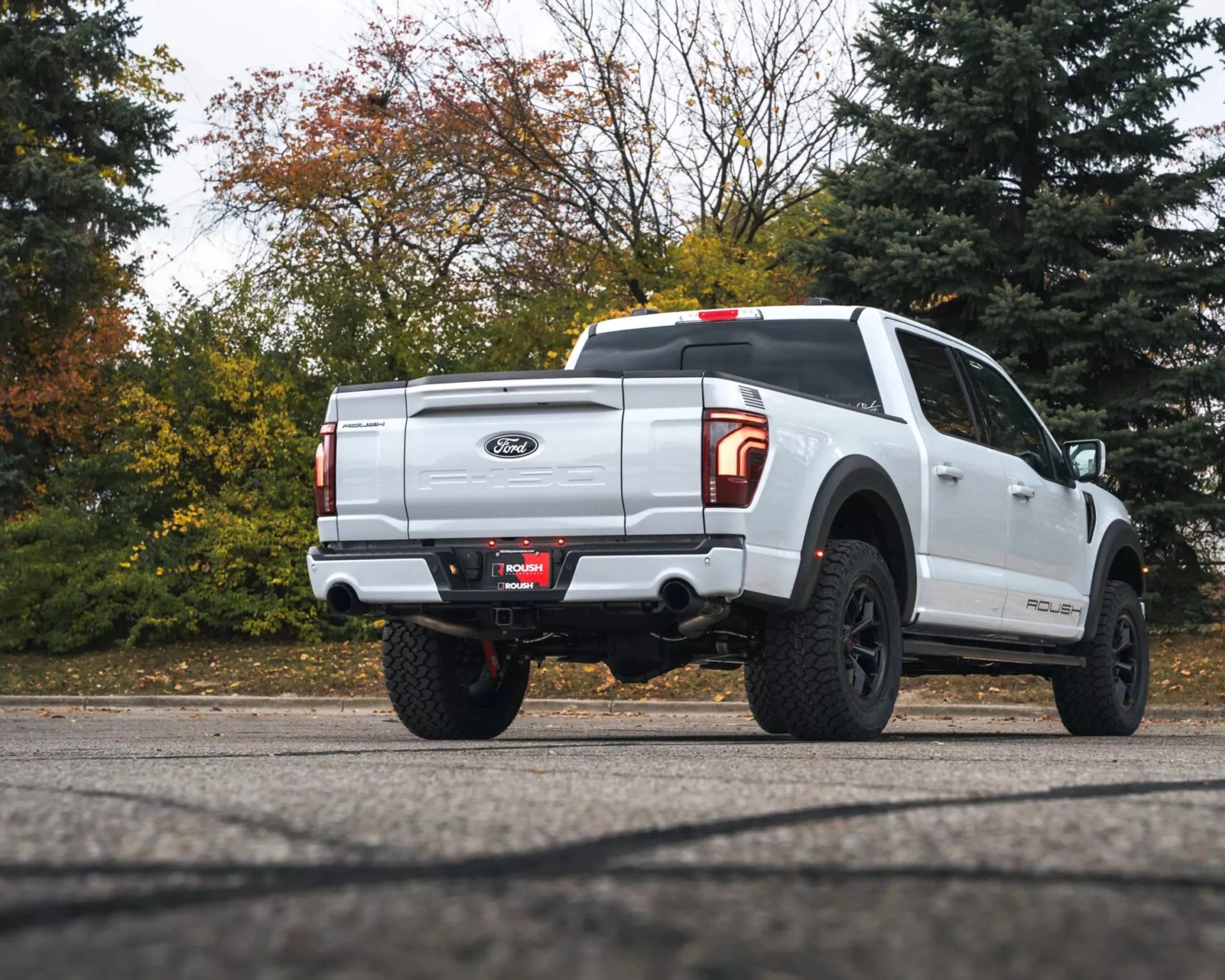 White Ford F-150 Roush pickup truck with black wheels parked on a paved surface with autumn trees in the background.