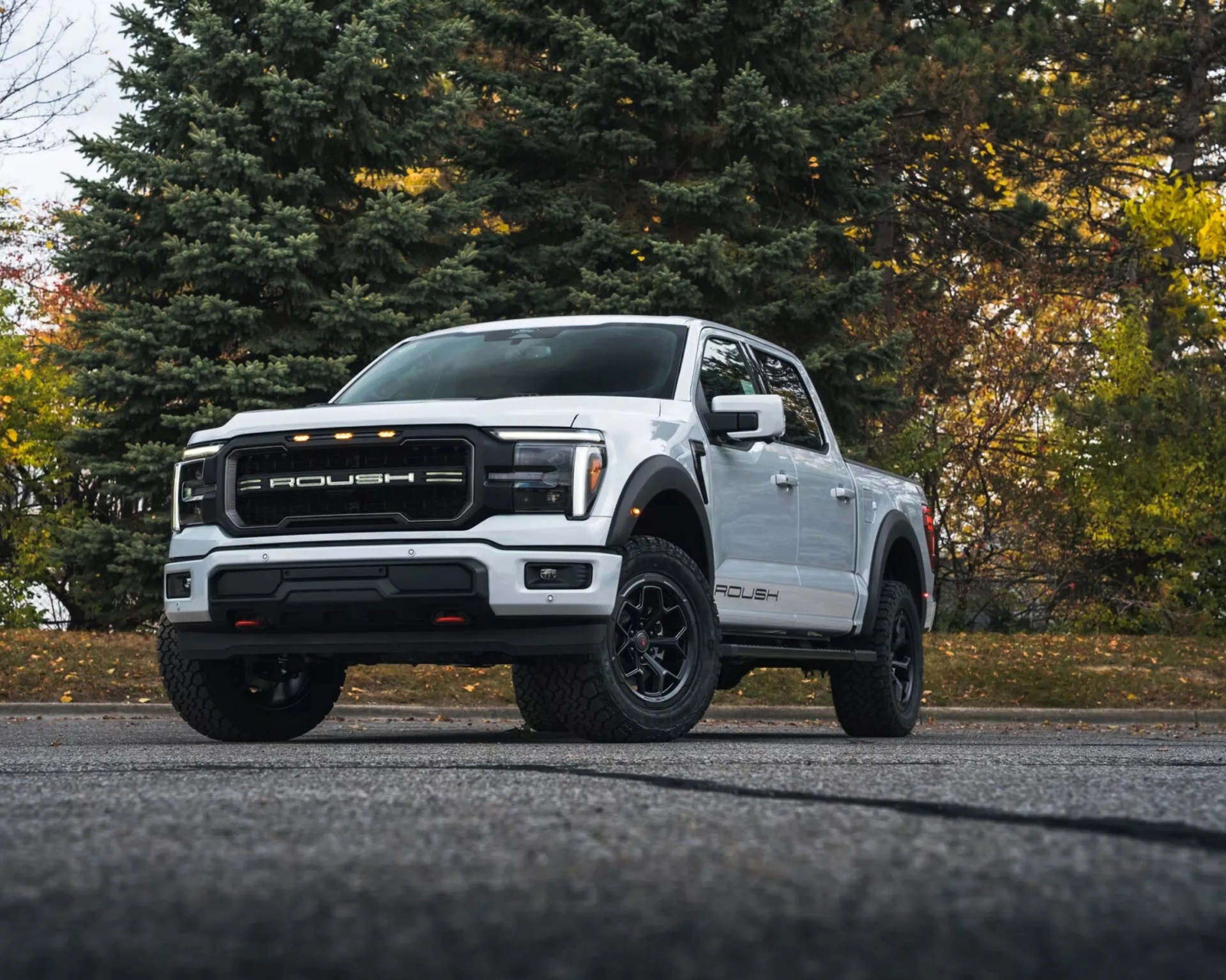 White Roush pickup truck with black grille and off-road tires parked on asphalt with trees in the background.