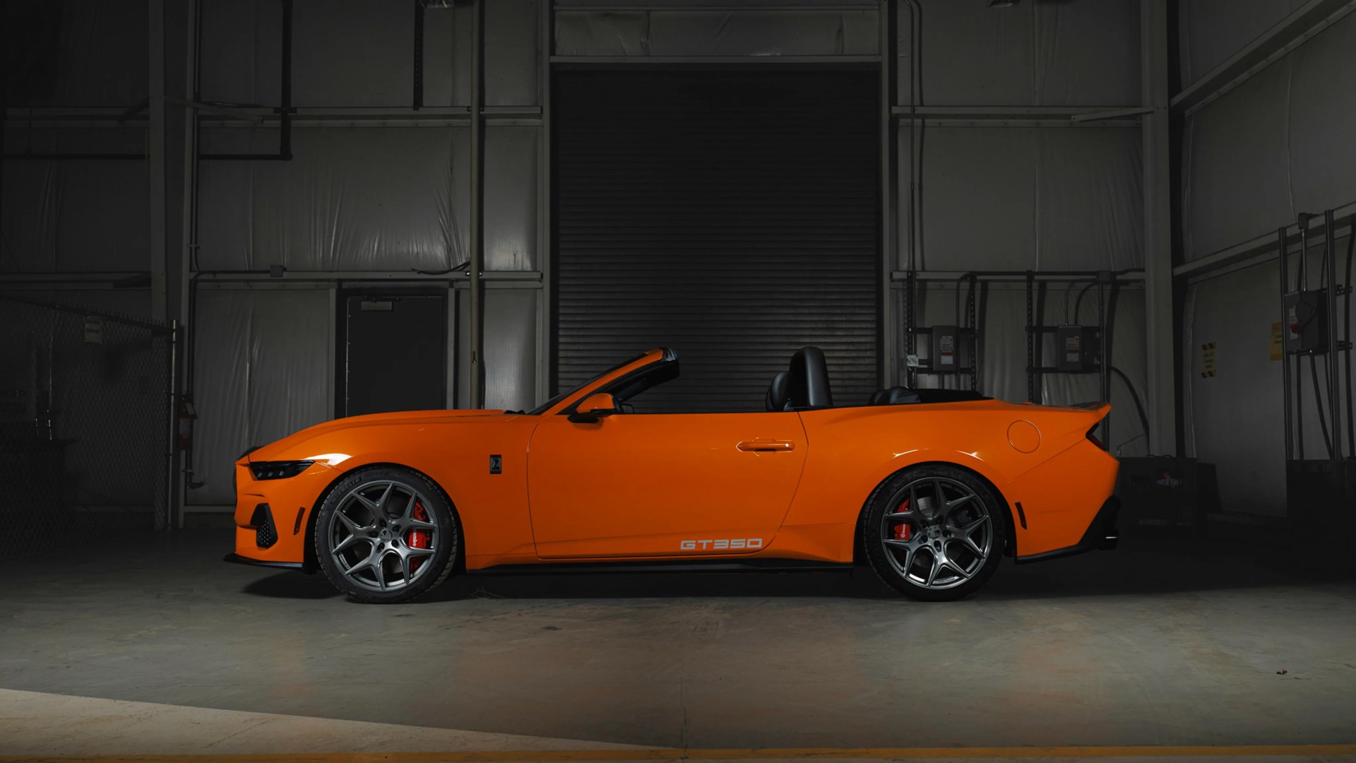 Orange Ford Mustang GT350 convertible with black interior parked indoors against a dark industrial backdrop.