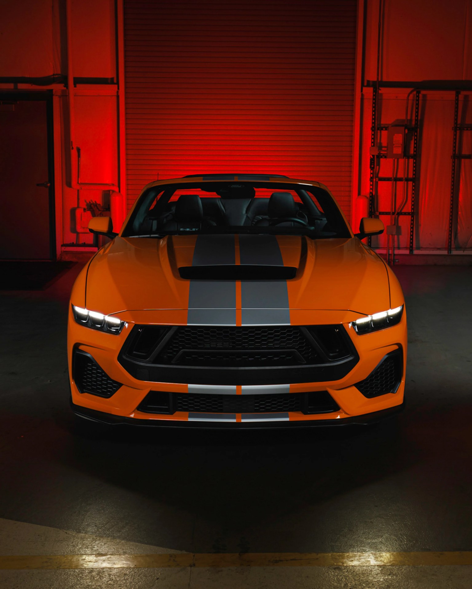 Orange sports car with black racing stripes and LED headlights in a dimly lit garage.