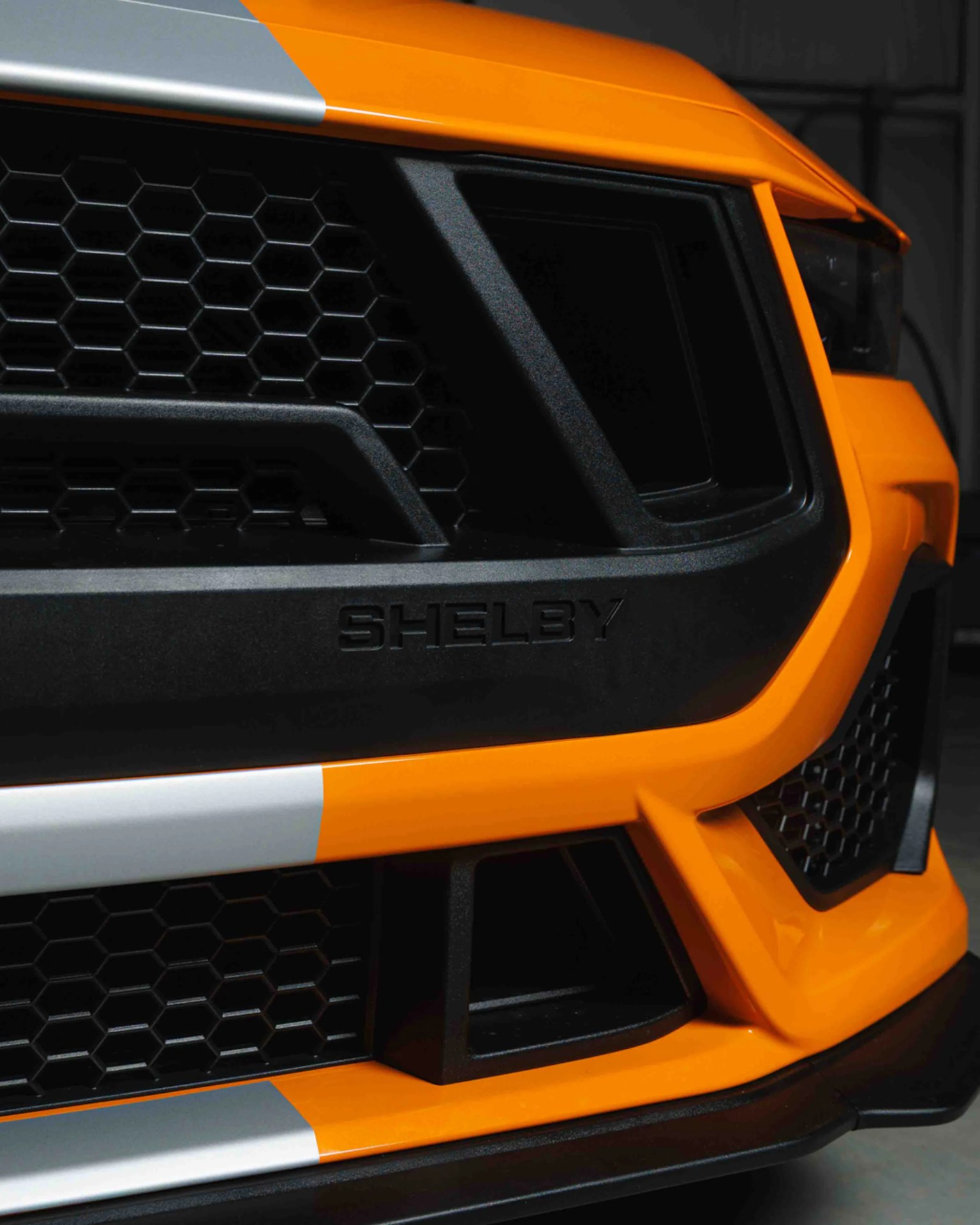 Close-up of the front grille of an orange Shelby car with black honeycomb mesh and silver racing stripes.