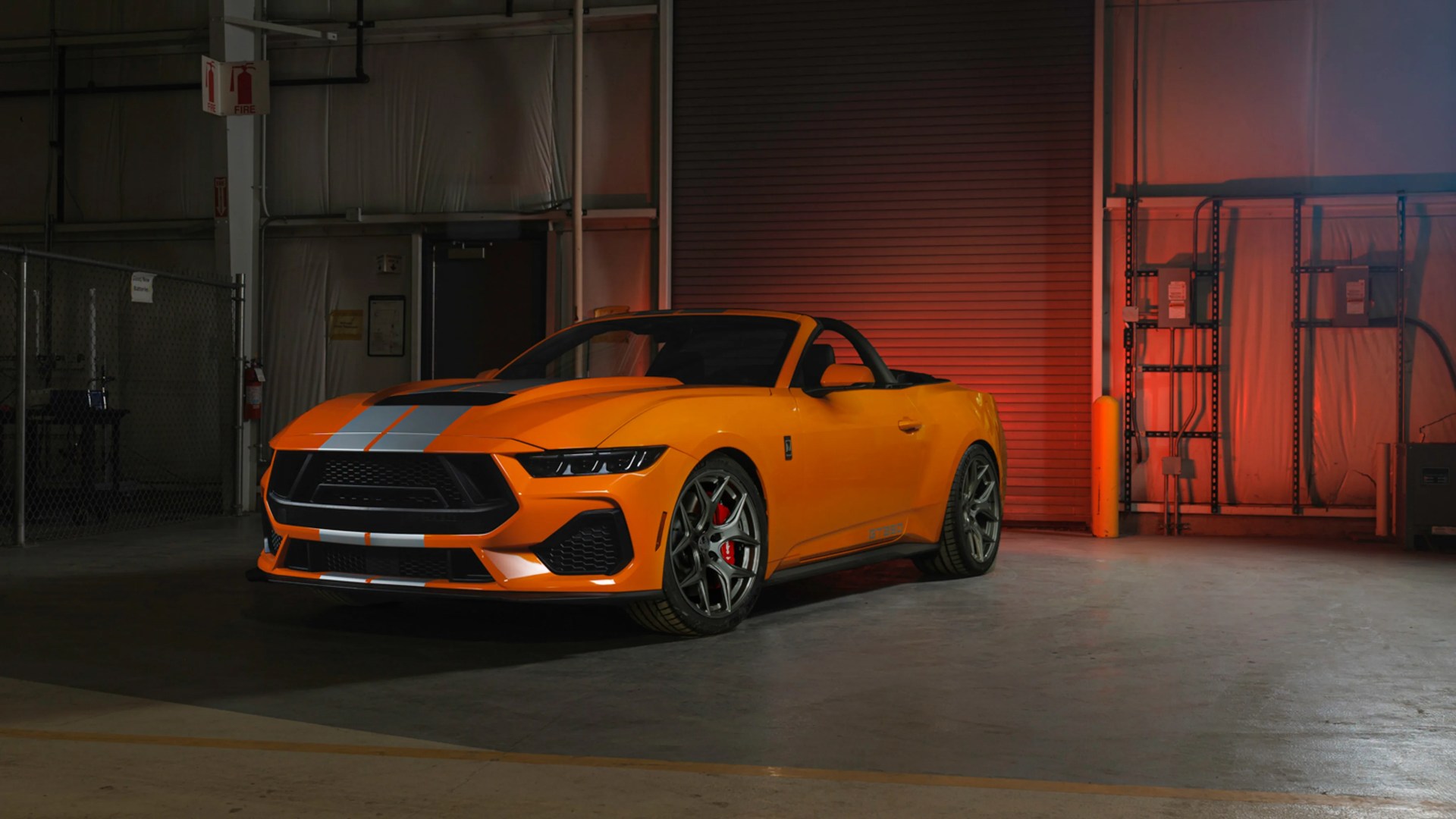 Orange convertible sports car with silver racing stripes and red brake calipers in a dimly lit garage.