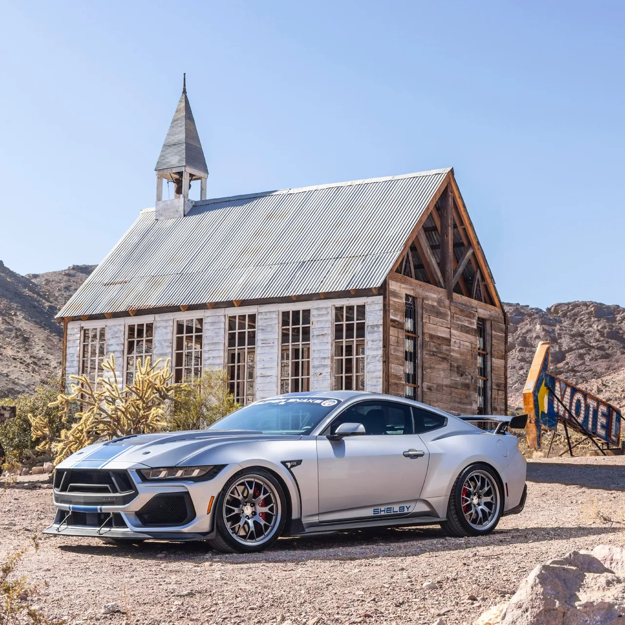 Silver Shelby sports car with blue racing stripes parked in front of a rustic wooden building with a metal roof.