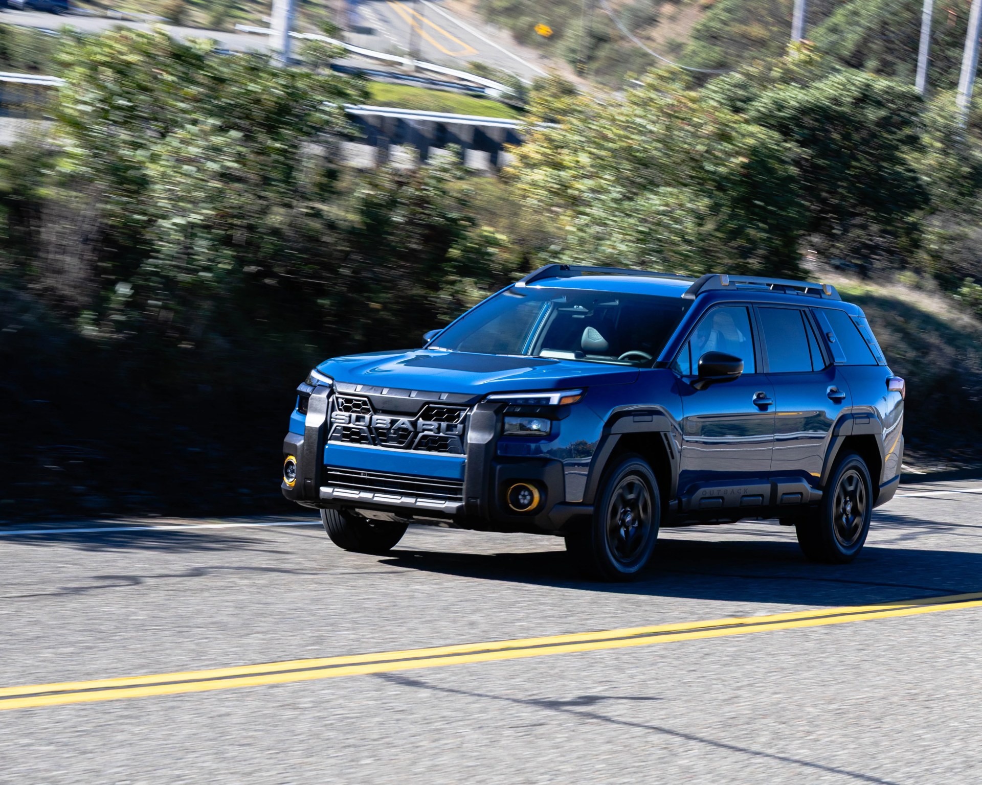 Blue Subaru SUV with black trim driving on a paved road with greenery in the background.