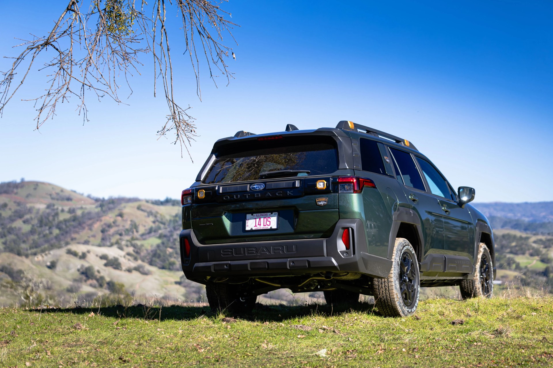 Dark green Subaru Outback SUV with black trim parked on grassy hill with mountains in the background.