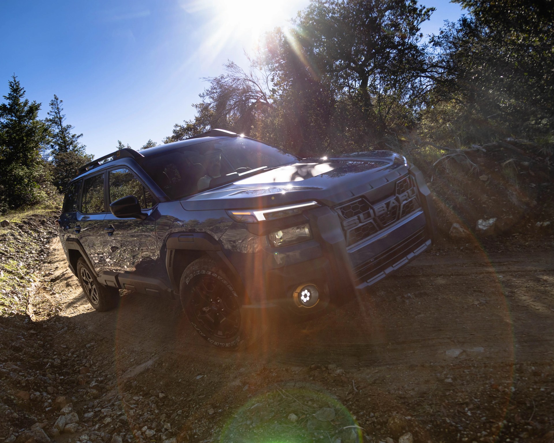 Dark gray Subaru SUV with black wheels on a dirt trail in a wooded area with sunlight glare.