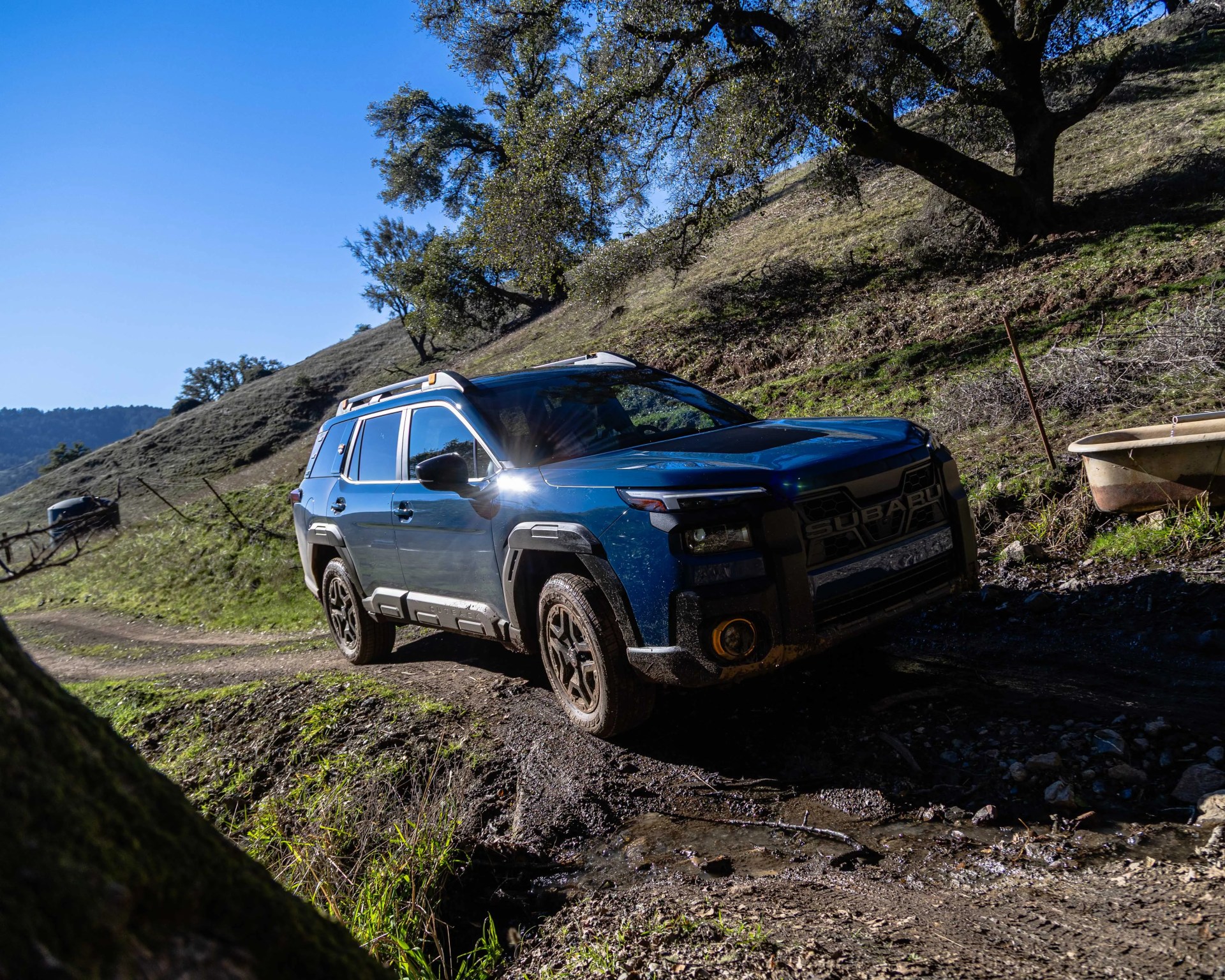 Blue Subaru SUV with roof rails driving on a muddy dirt road in a hilly, grassy landscape.