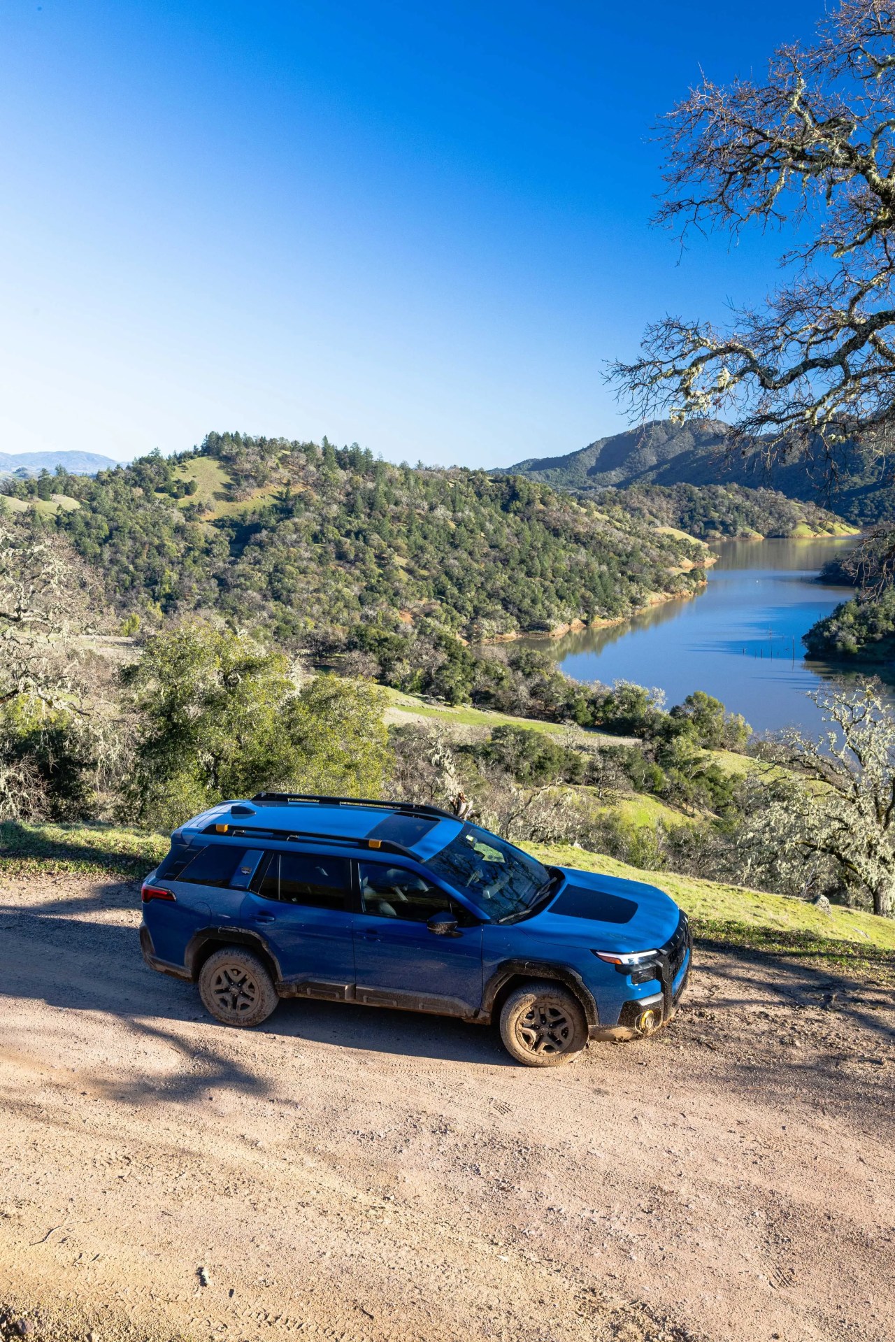 Blue SUV with black roof rack parked on a dirt road overlooking a lake and green hills.