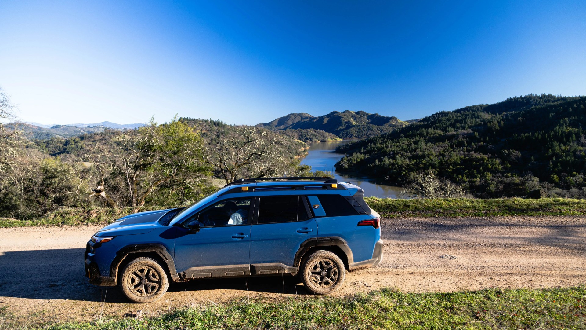 Blue SUV with roof rails parked on a dirt road overlooking a river and forested hills.