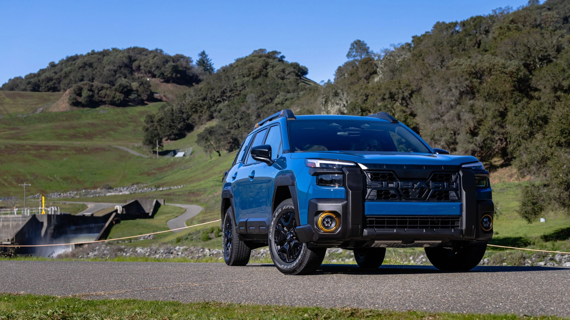 Blue Subaru SUV with black trim and yellow fog light rings parked on a paved road with green hills and trees in the background.