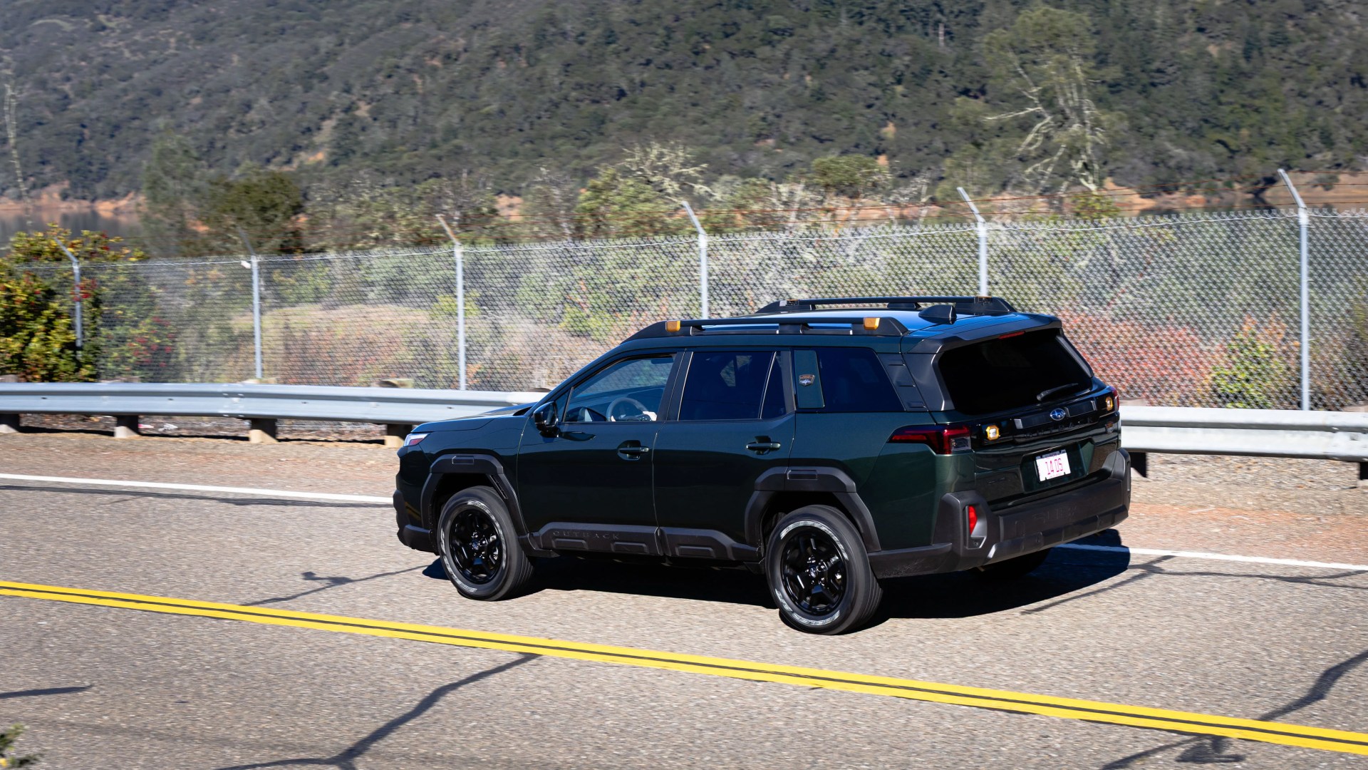 Dark green Subaru Outback SUV with black wheels driving on a paved road near a chain-link fence.