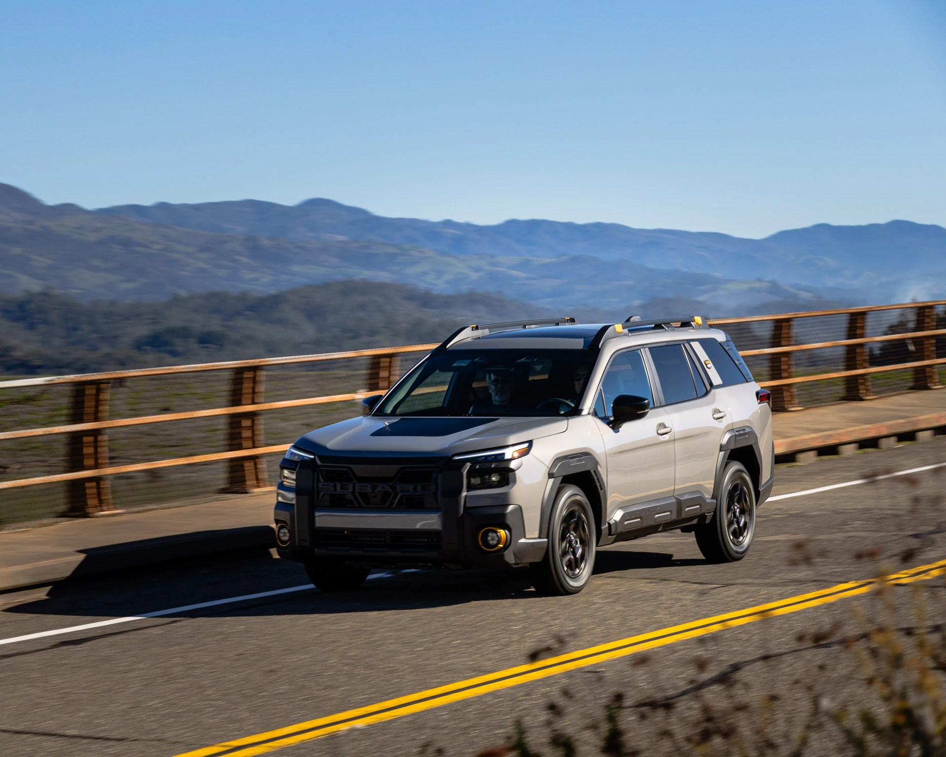 Silver Subaru SUV with black accents driving on a mountain road with wooden guardrails.