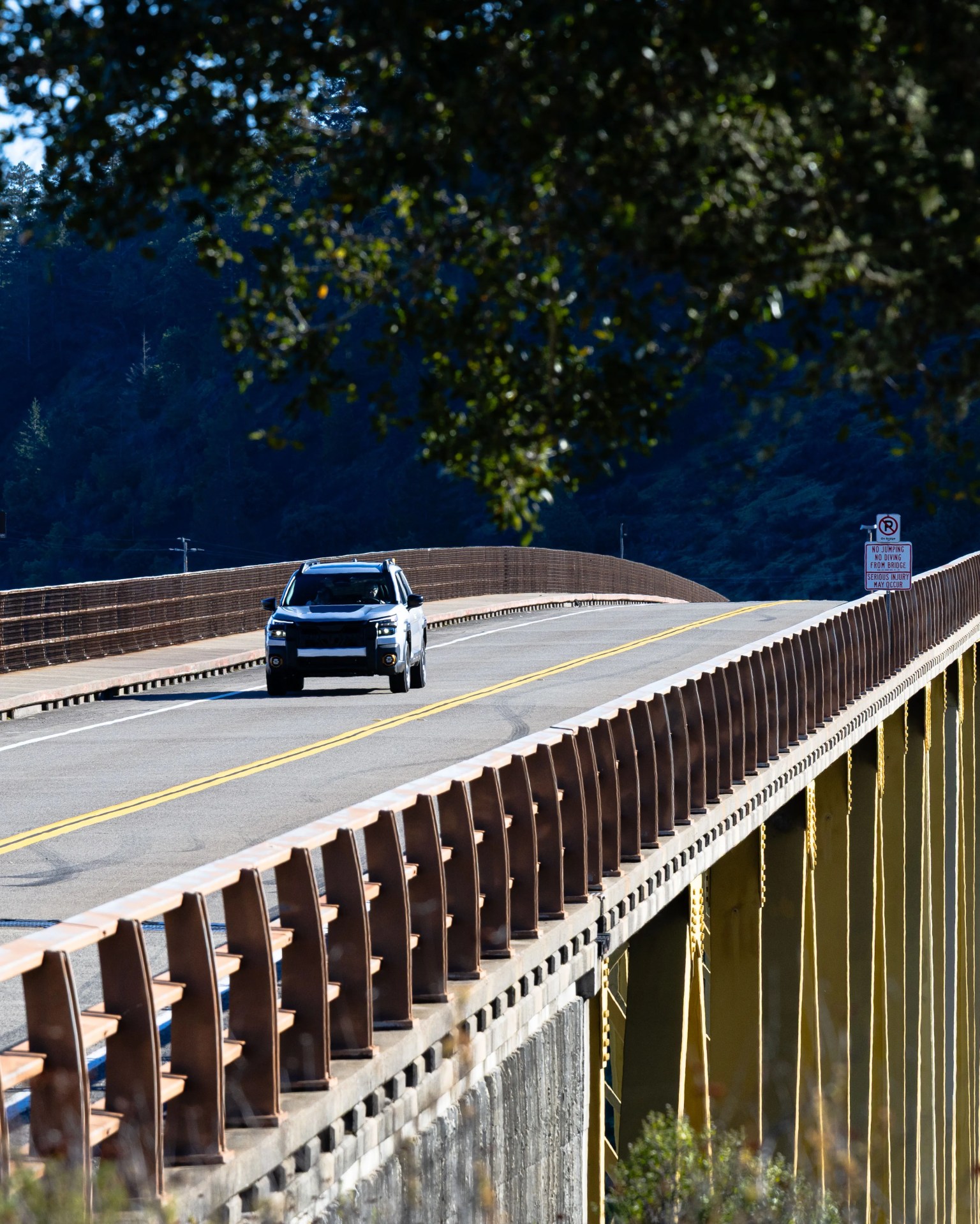 Black SUV driving on a bridge with brown railings and yellow support beams under a tree canopy.