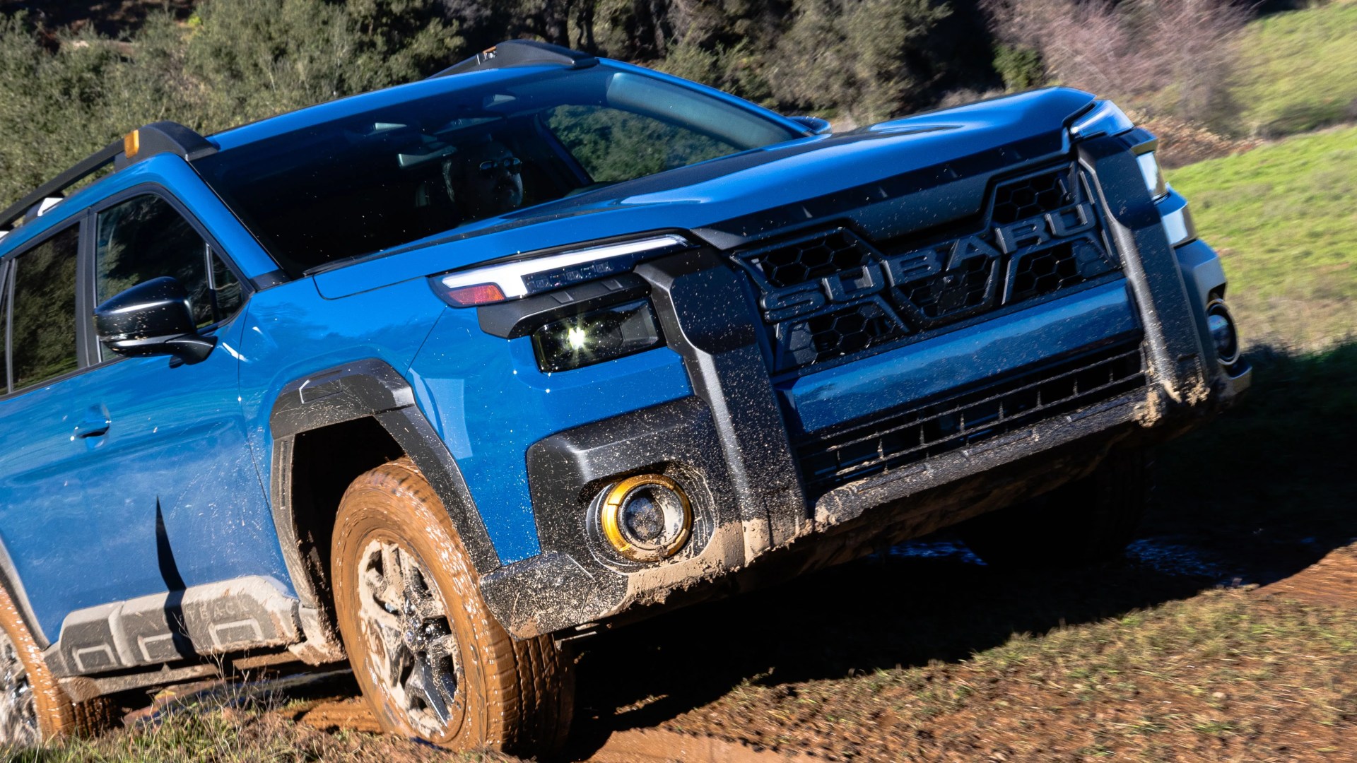 Blue Subaru SUV with mud-covered tires driving off-road on a grassy dirt path.