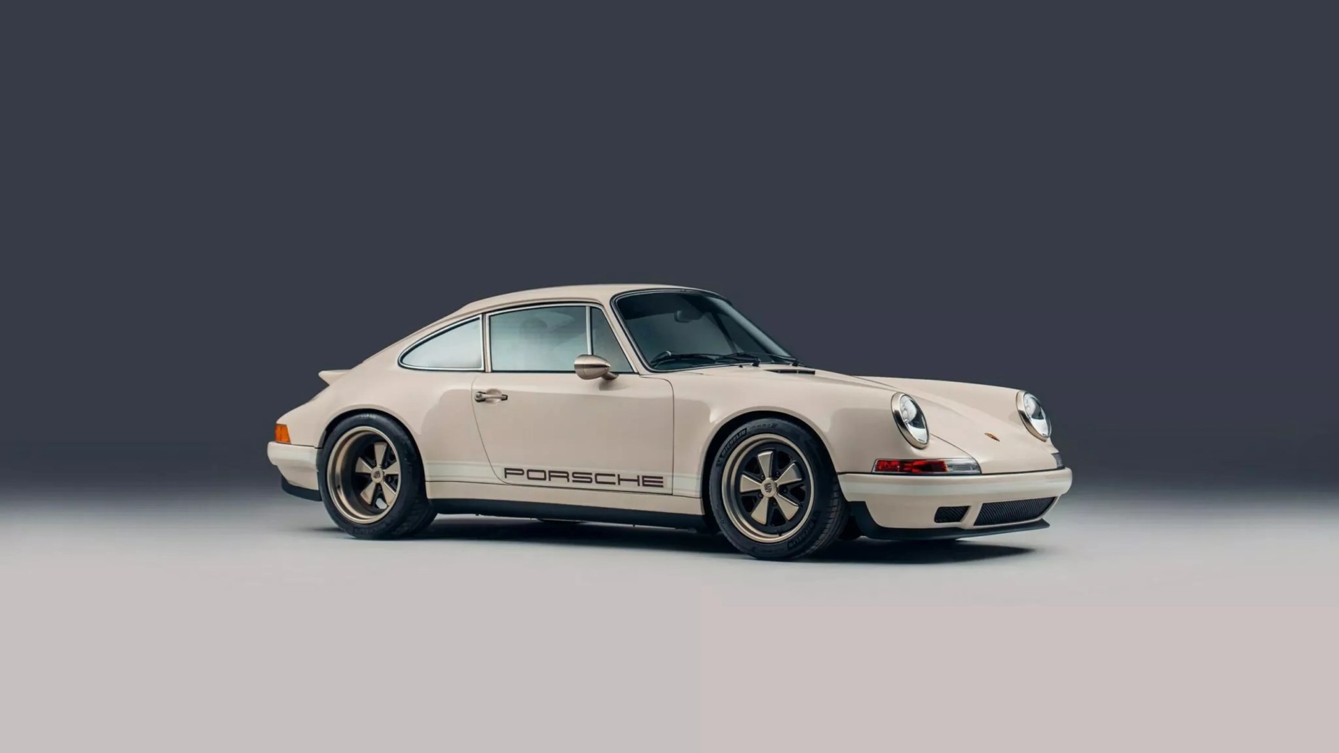 Cream-colored classic Porsche 911 with black and gold wheels on a two-tone gray background.