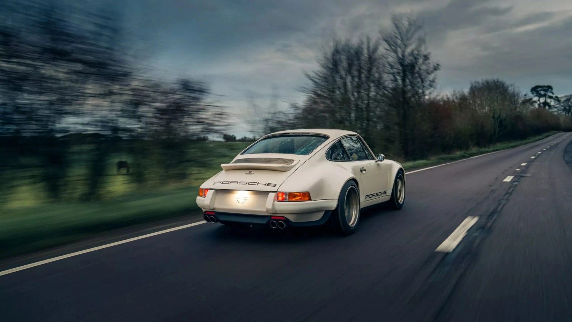 Cream-colored Porsche 911 with wide tires driving on a rural road with blurred trees.