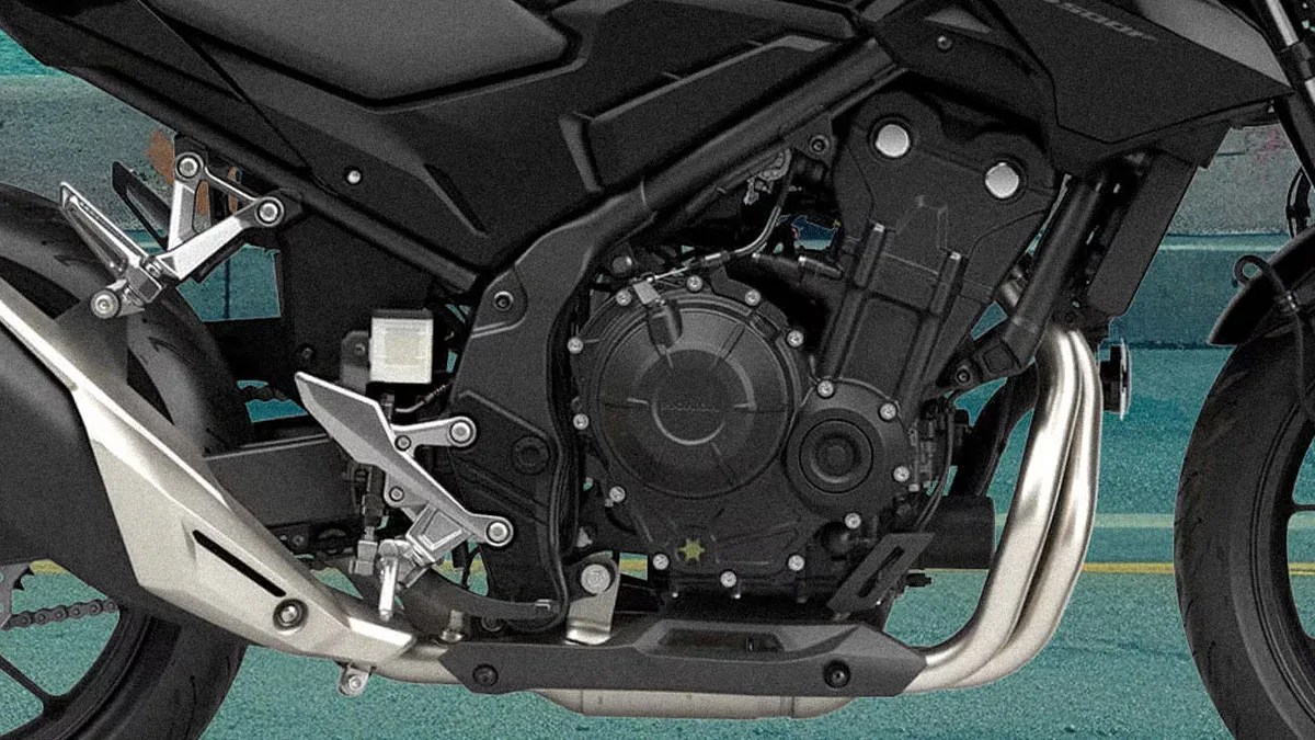 Close-up of a black motorcycle engine with silver exhaust pipes and foot pegs on a teal pavement background.