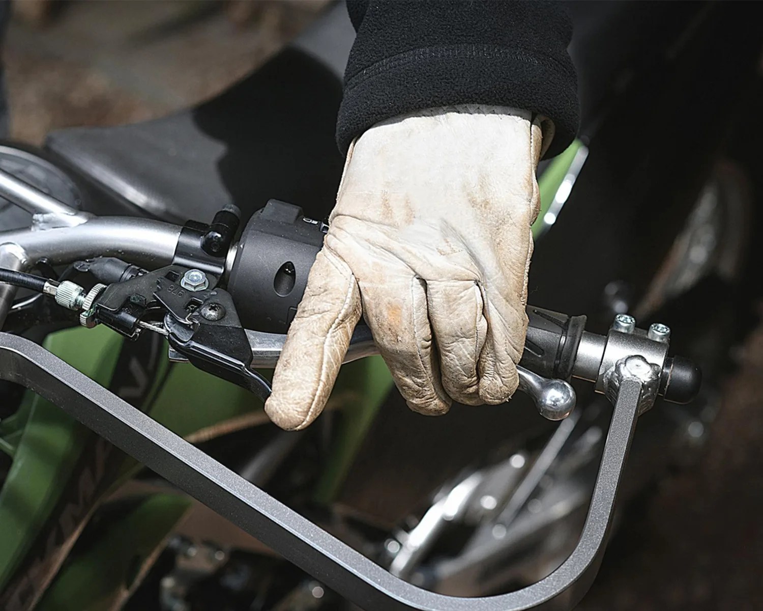 Hand wearing a worn beige leather glove gripping a motorcycle handlebar with a brake lever and handguard.