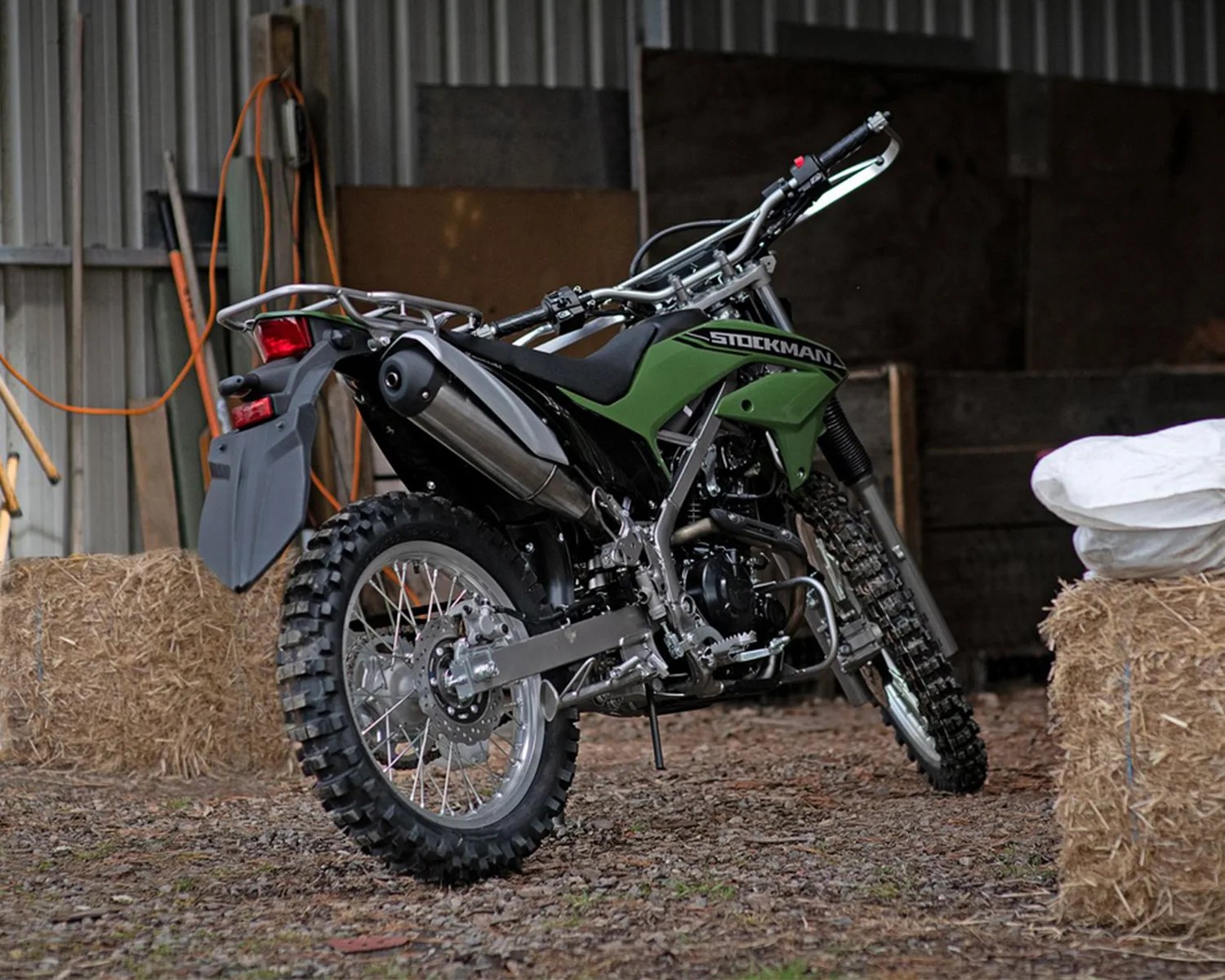 Green and black Stockman dirt bike with knobby tires parked on gravel inside a barn with hay bales nearby.