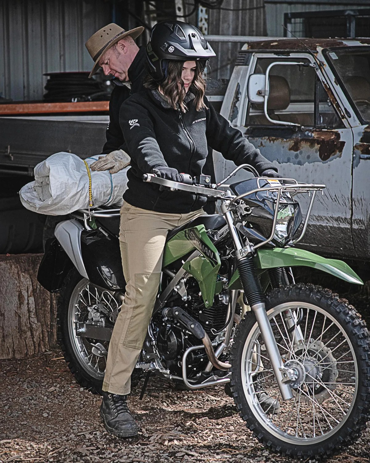 Person wearing a black helmet and jacket sitting on a green and black off-road motorcycle with a rolled tarp secured on the back.