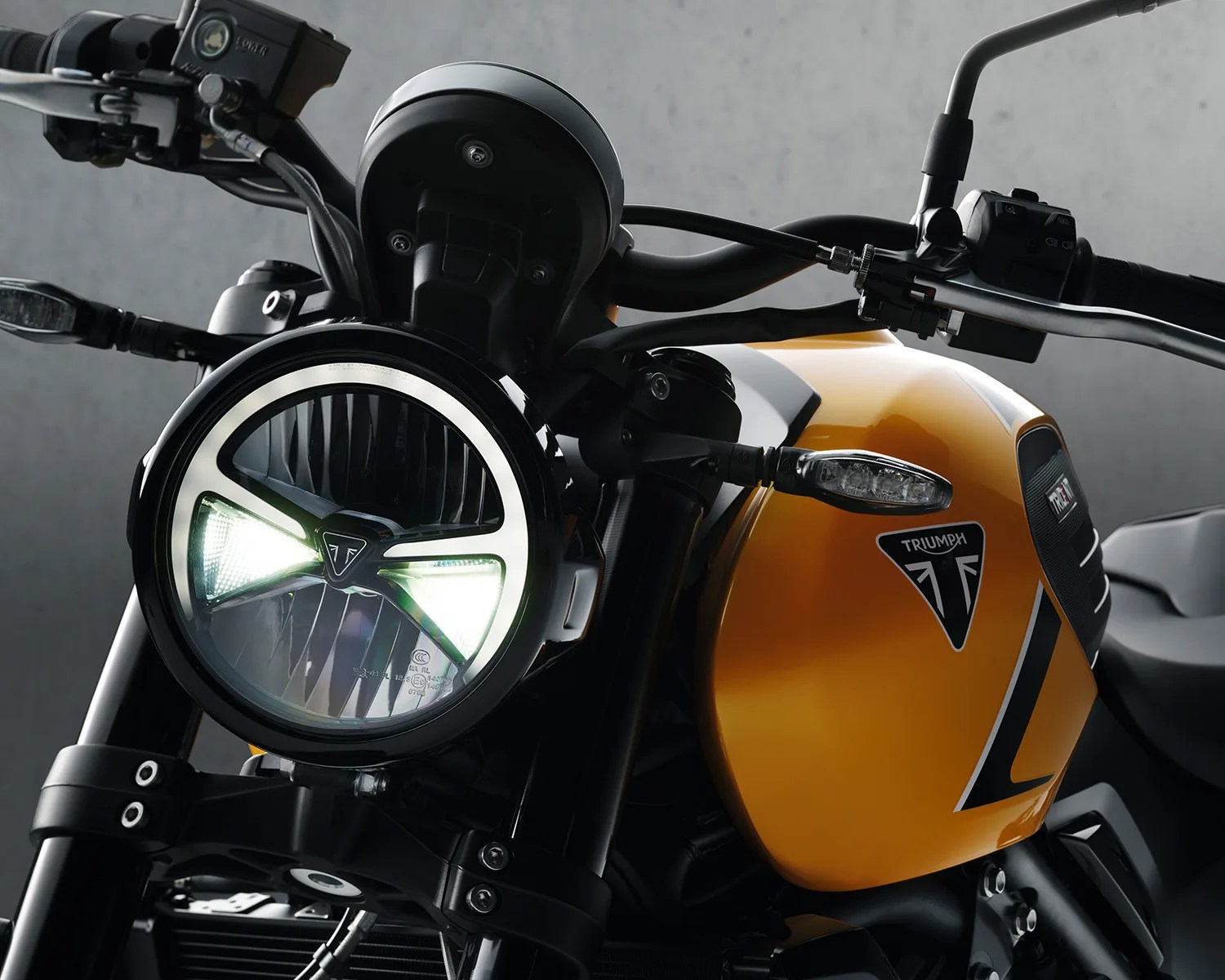 Close-up of a yellow Triumph motorcycle fuel tank and illuminated round headlight with black handlebars.