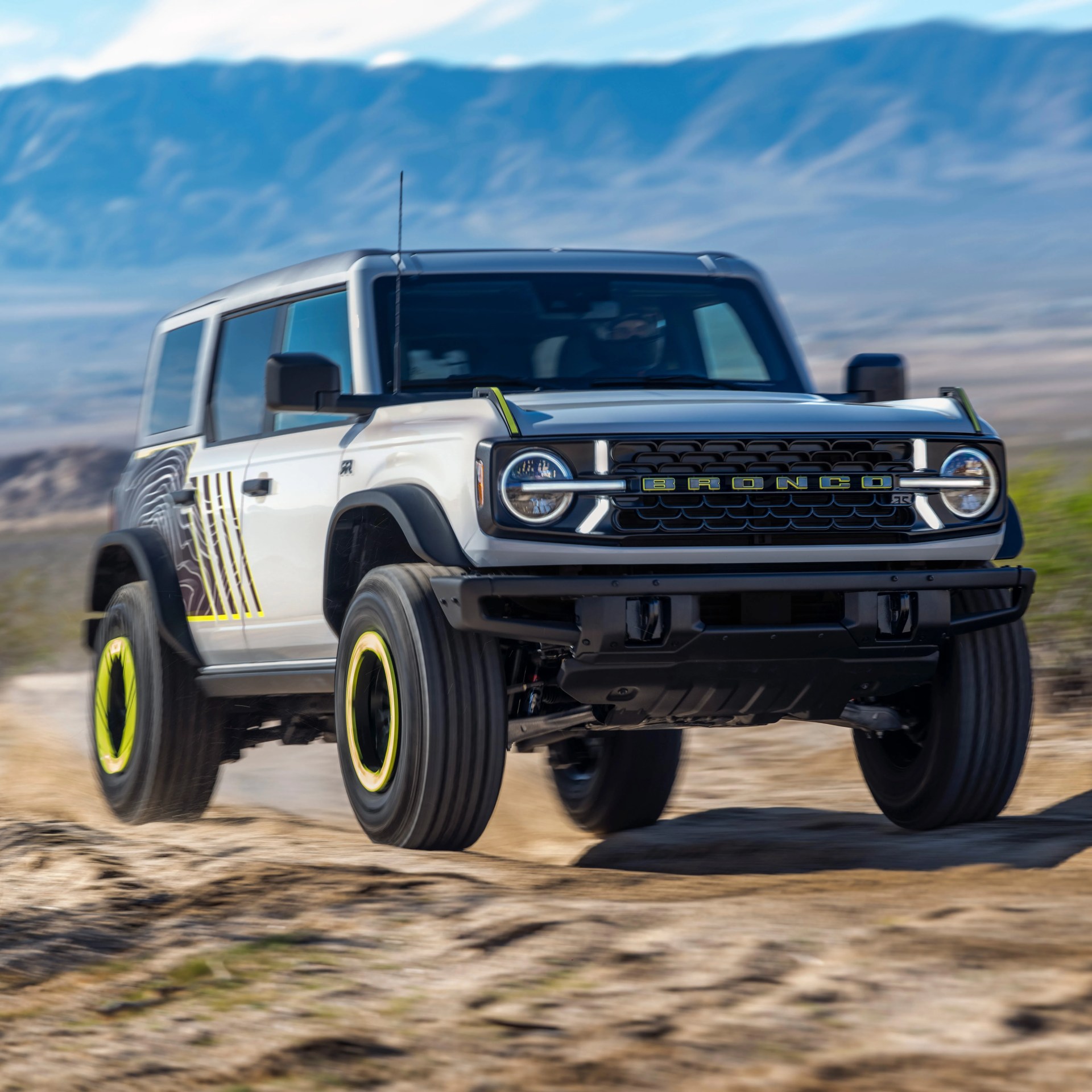 White Ford Bronco SUV with black and yellow accents driving off-road on a dirt trail with mountains in the background.