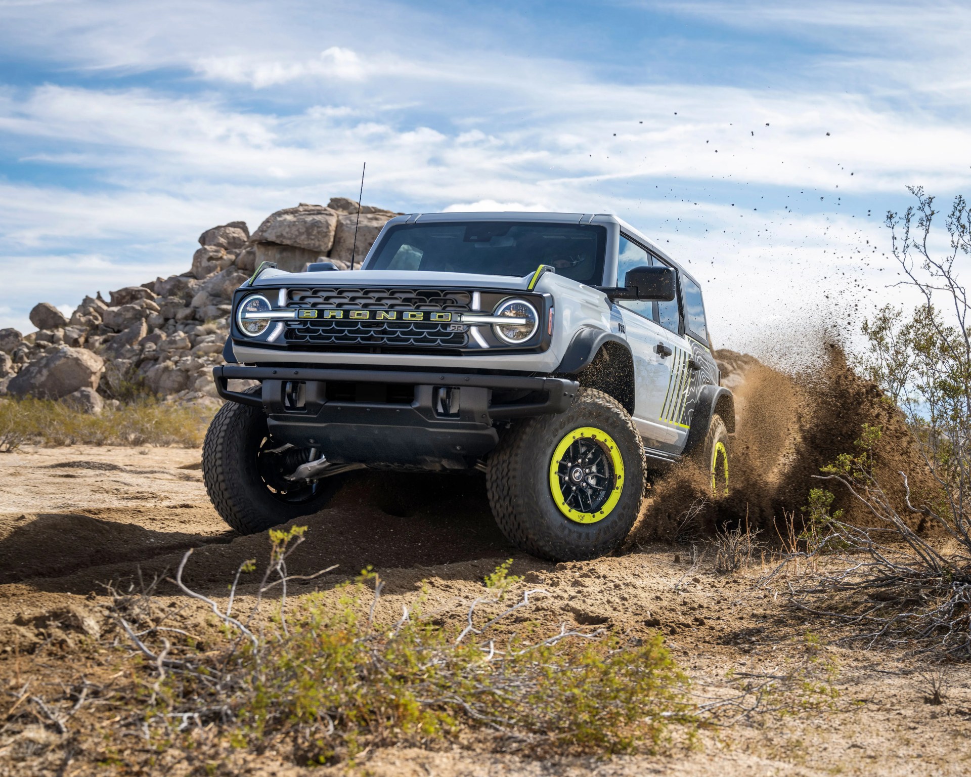 Silver Ford Bronco with neon yellow-accented wheels kicking up dirt in a desert landscape.