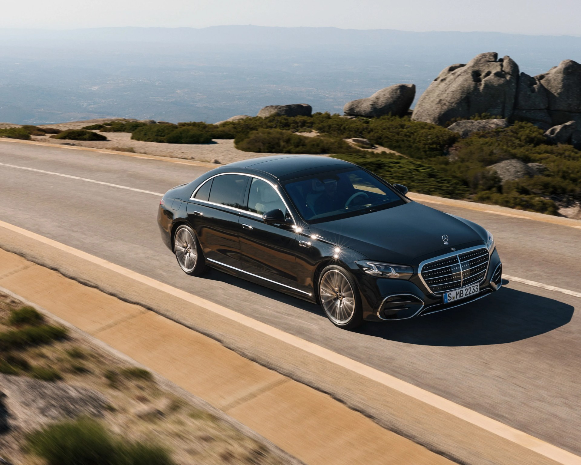 Black Mercedes-Benz luxury sedan driving on a mountain road with rocky terrain in the background.