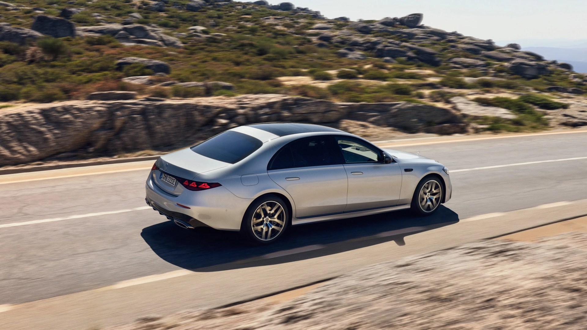 Silver Mercedes-Benz sedan driving on a mountain road with rocky terrain in the background.