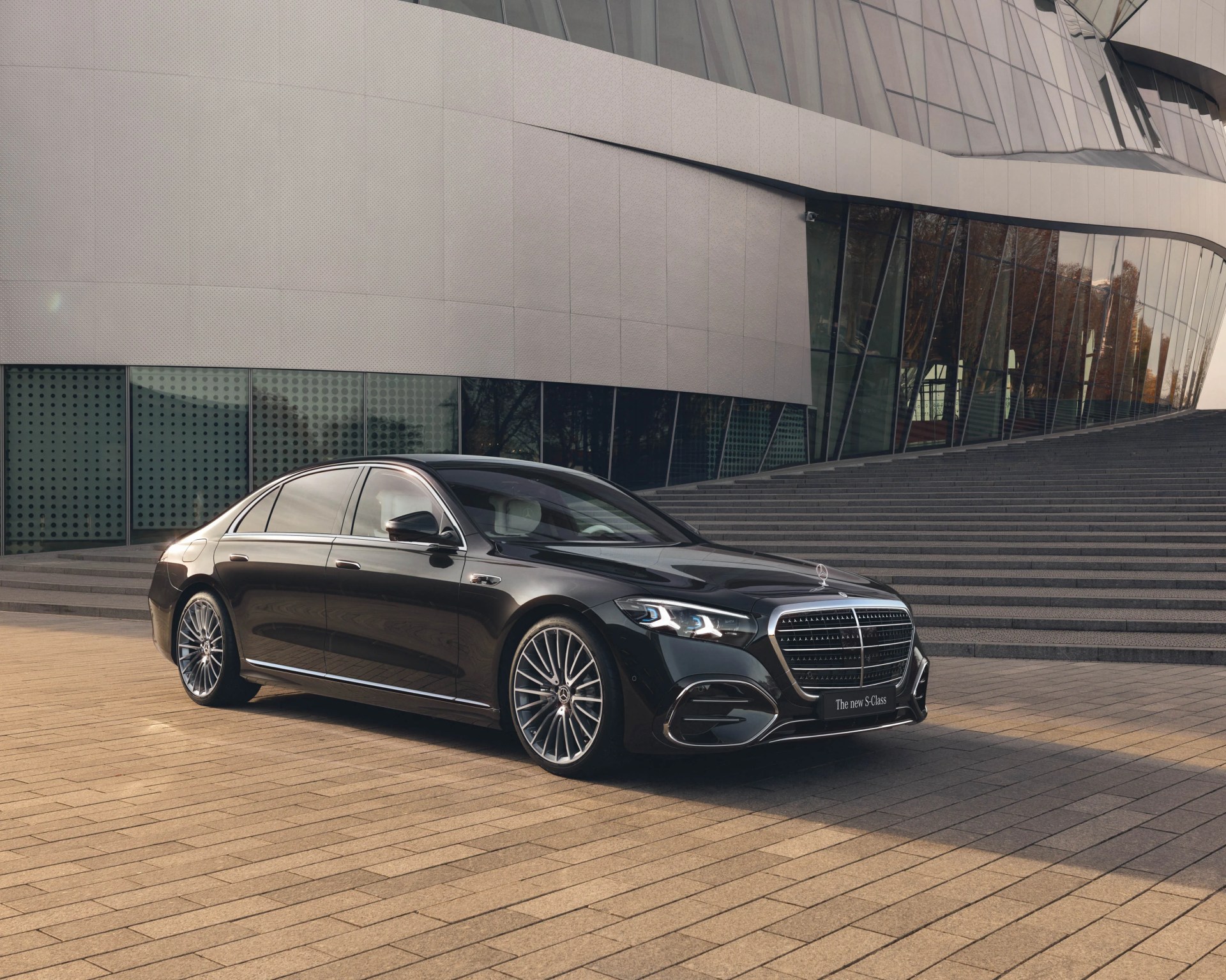 Black Mercedes-Benz S-Class sedan parked on a paved area in front of a modern building with large glass windows and stairs.
