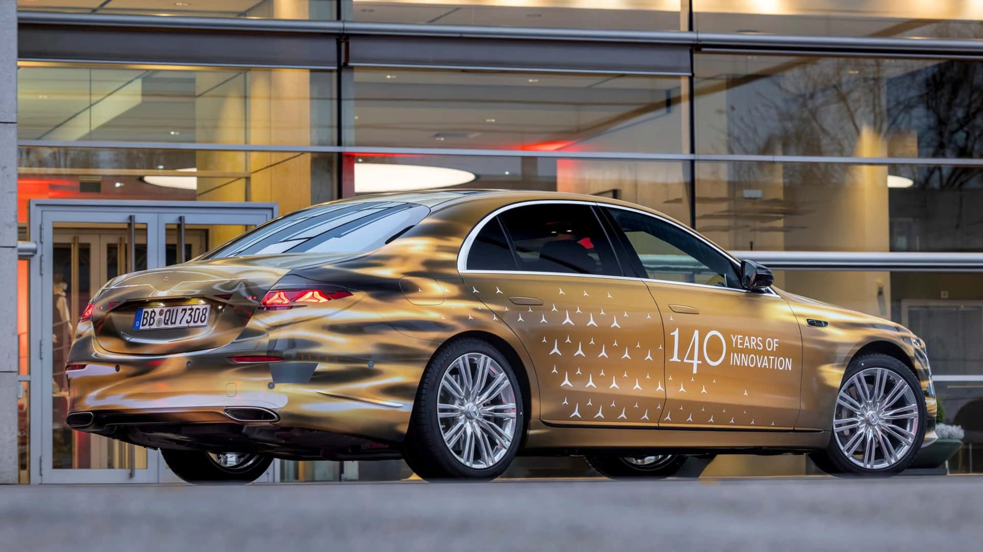 Gold and black luxury sedan with "140 Years of Innovation" text and Mercedes logos on the side, parked in front of a glass building.