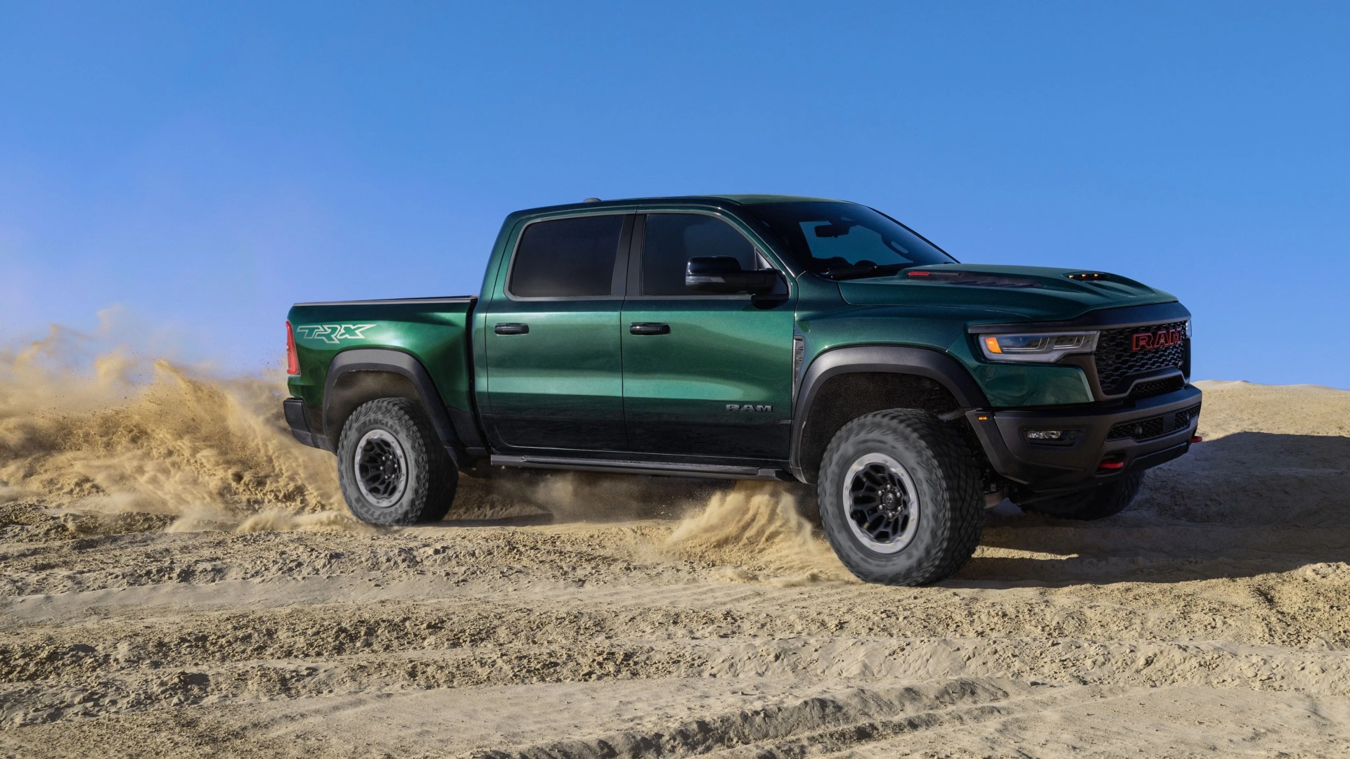 Green RAM TRX pickup truck kicking up sand while driving on a sandy terrain under a clear blue sky.