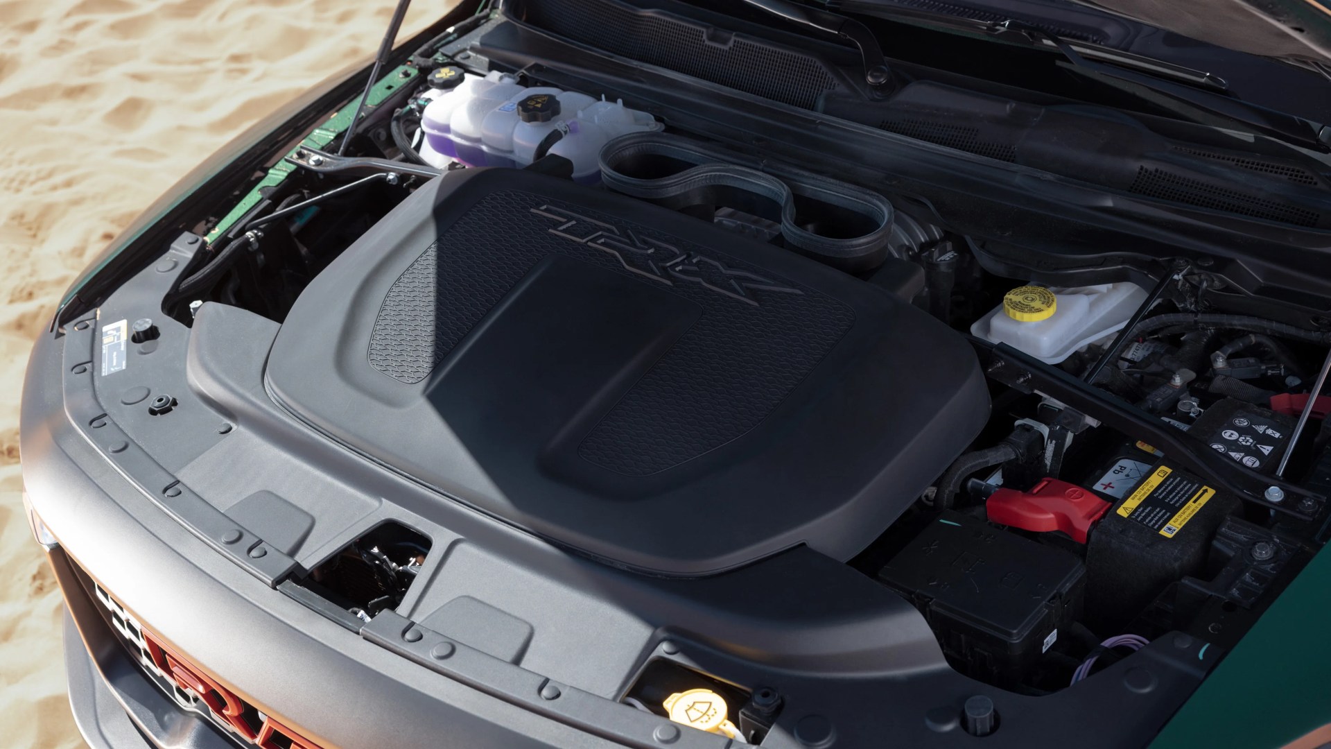 Engine compartment of a vehicle with a large black plastic engine cover labeled "TRX" and visible battery and fluid reservoirs.