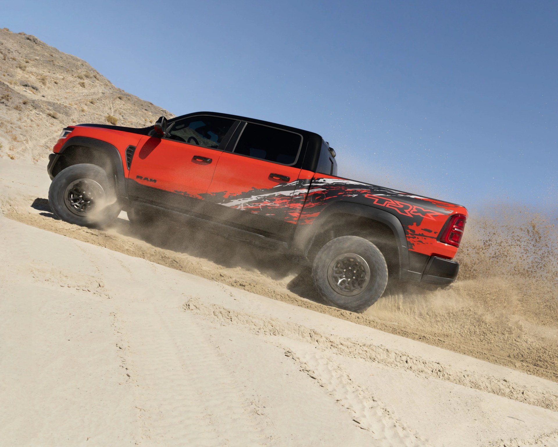 Red and black Ram TRX pickup truck kicking up sand while driving uphill on a sandy terrain.