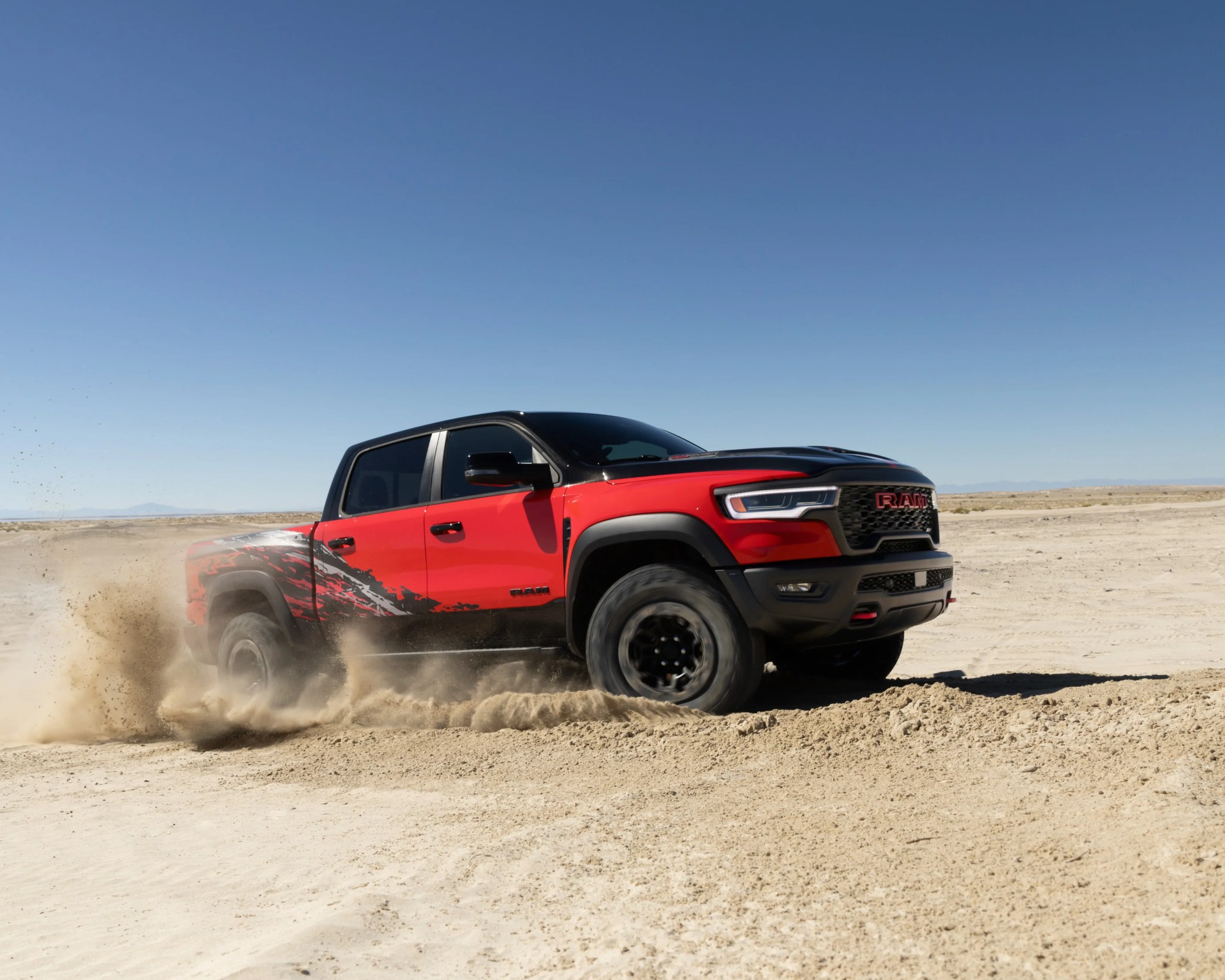 Red and black RAM pickup truck kicking up sand while driving on a desert terrain under a clear blue sky.