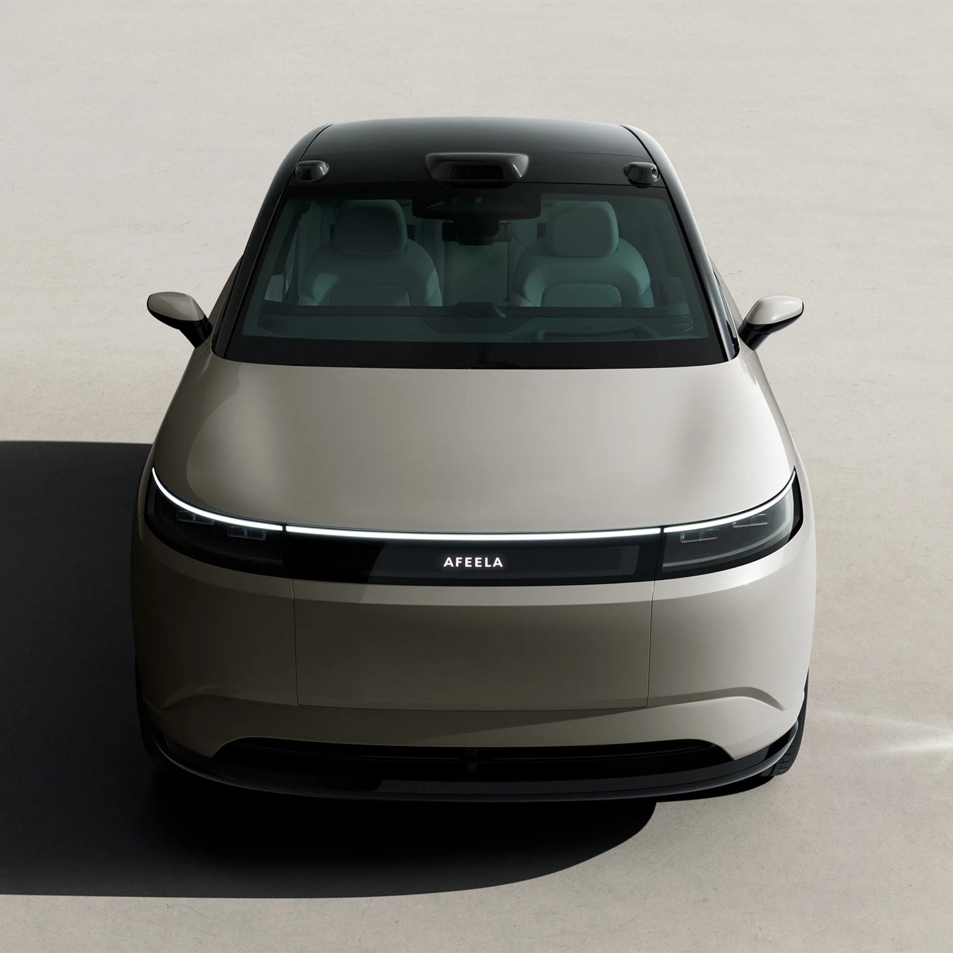 Front view of a beige Afeela electric car with a black roof and illuminated logo on a light gray surface.