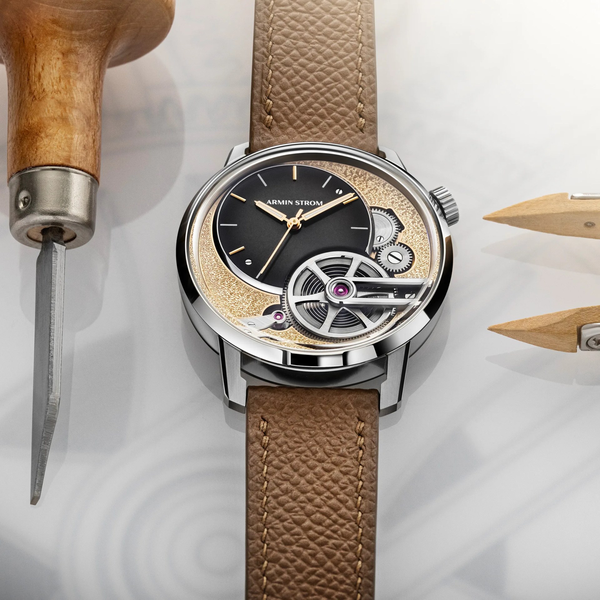 Silver Armin Strom wristwatch with black and gold textured dial, visible gears, and brown leather strap on a white surface.