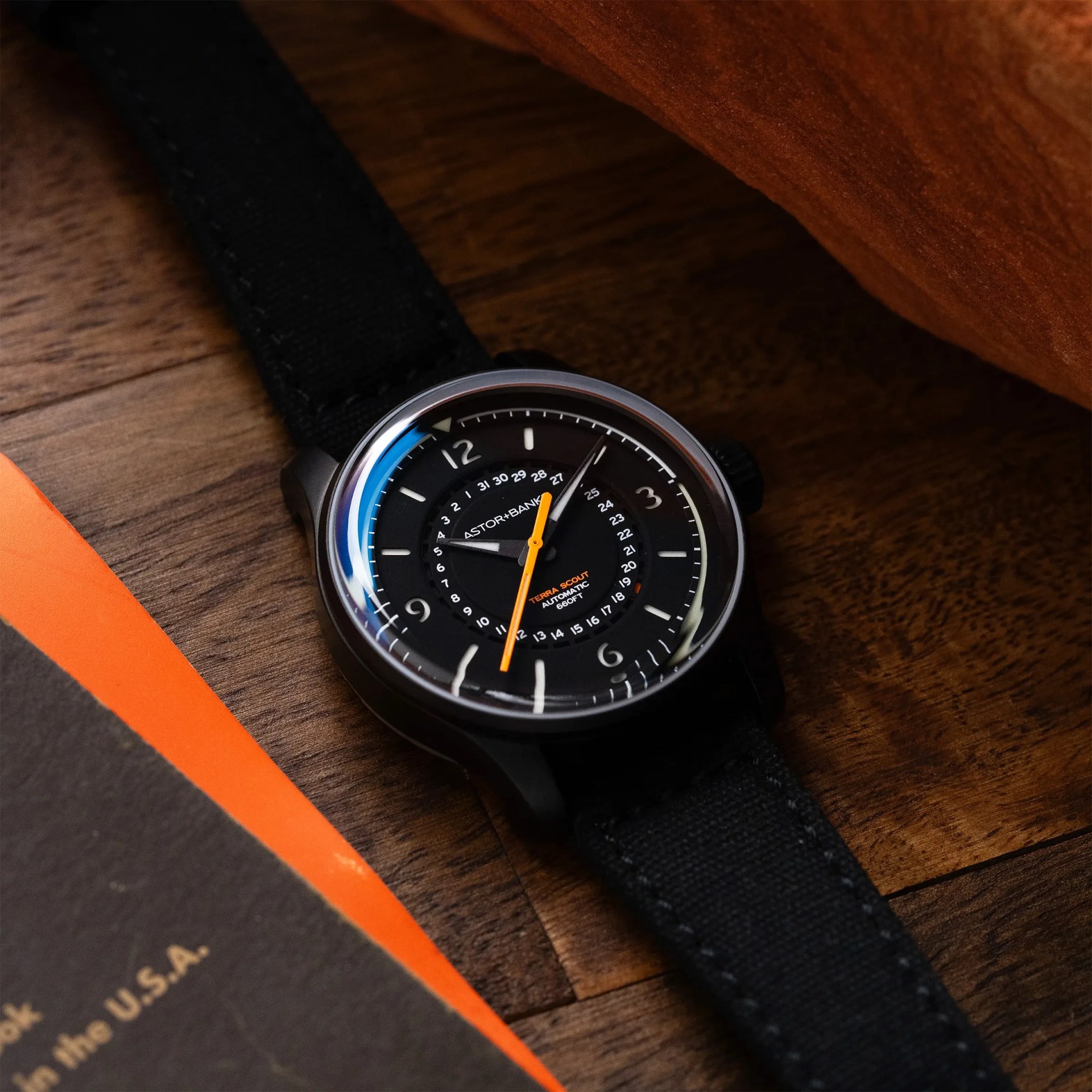 Black wristwatch with orange second hand and black fabric strap on a wooden surface next to an orange and black book.