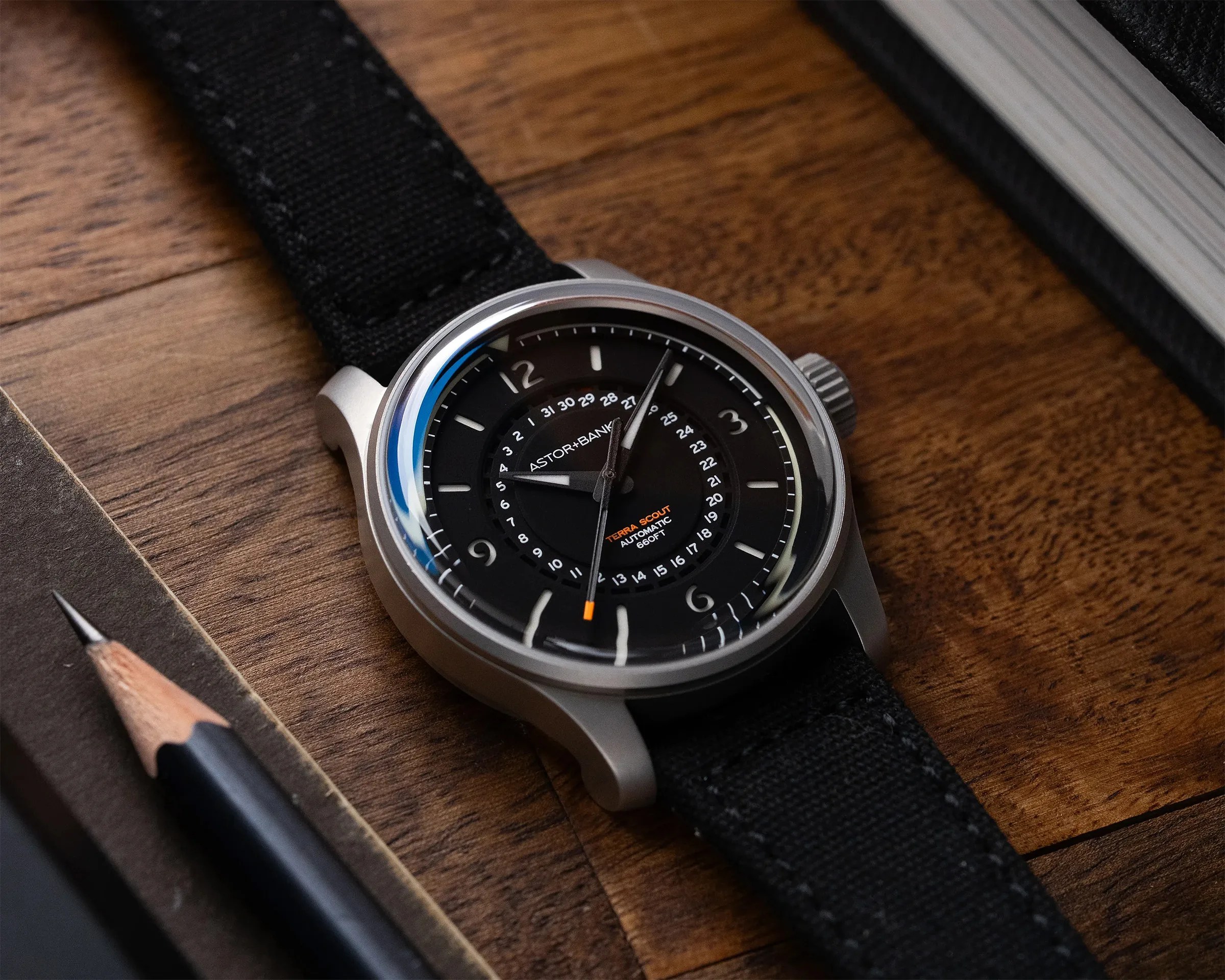 Black dial wristwatch with silver case and black fabric strap on a wooden surface next to a pencil and notebook.