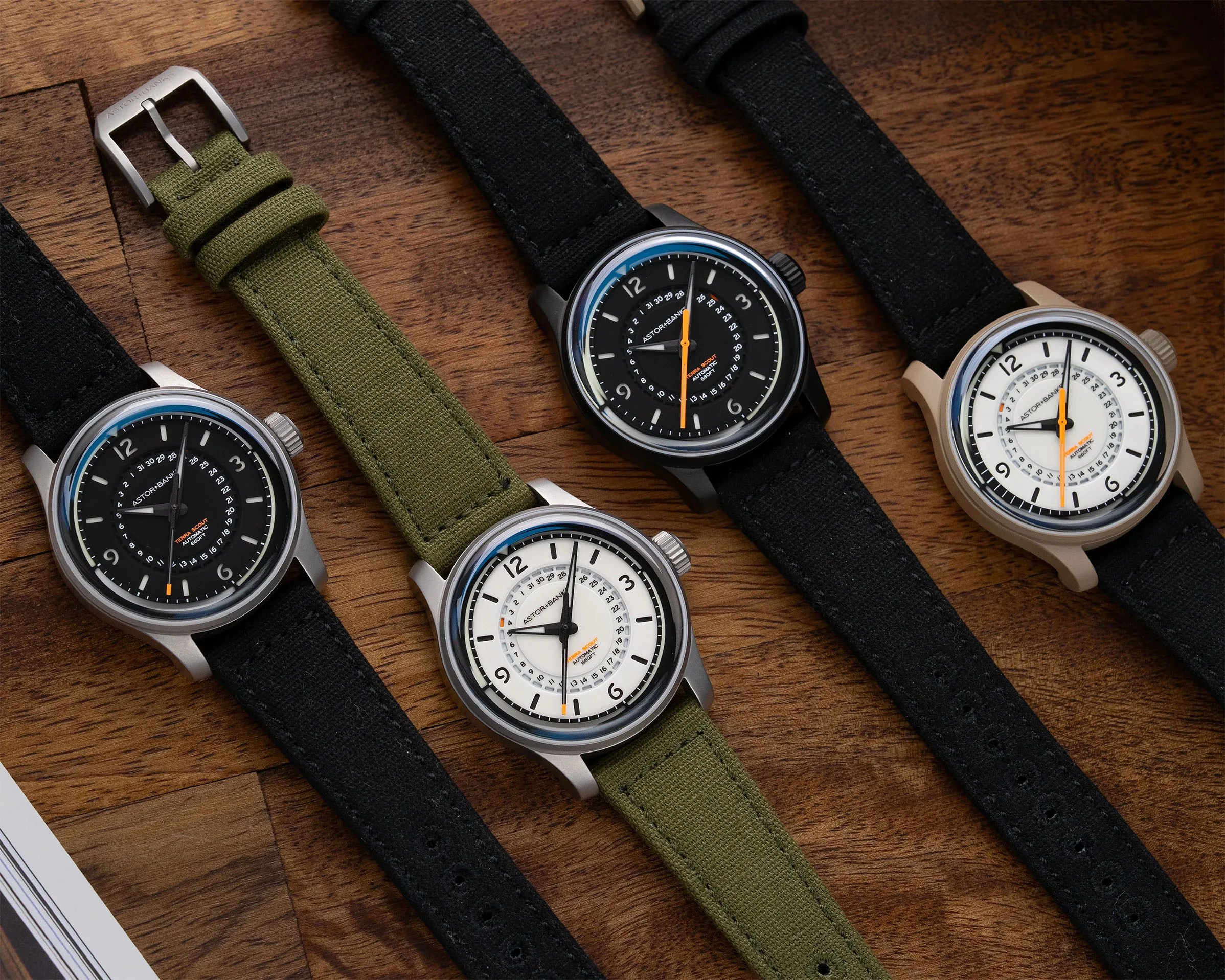 Four wristwatches with black or white dials, orange second hands, and black or green fabric straps on a wooden surface.