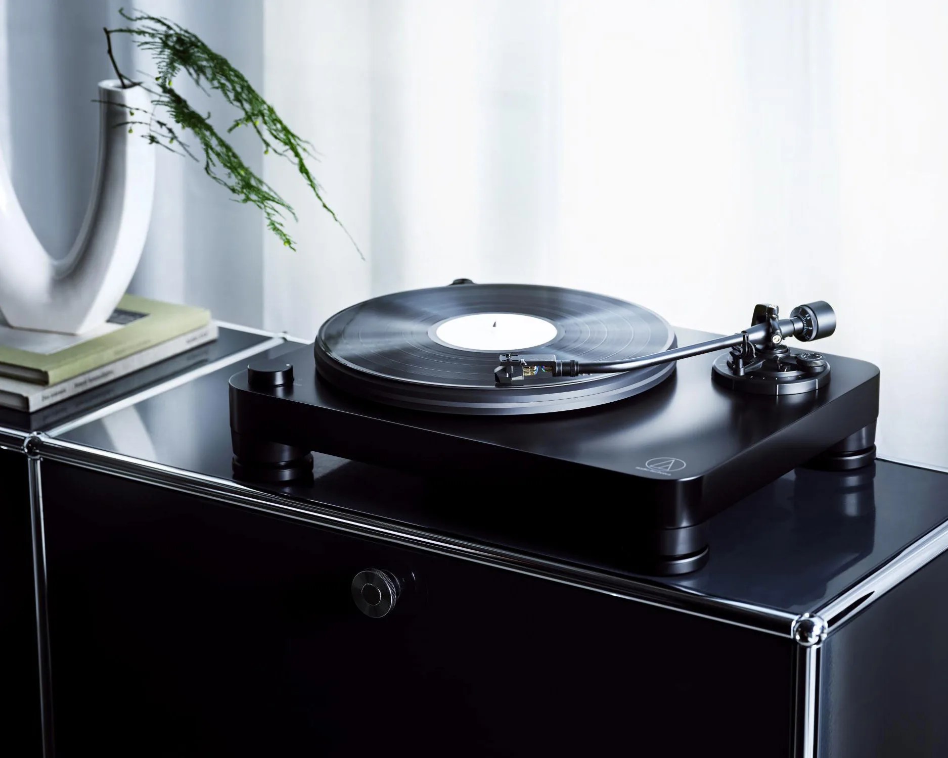 Black modern turntable with a vinyl record on a glossy black cabinet next to a white vase and books.