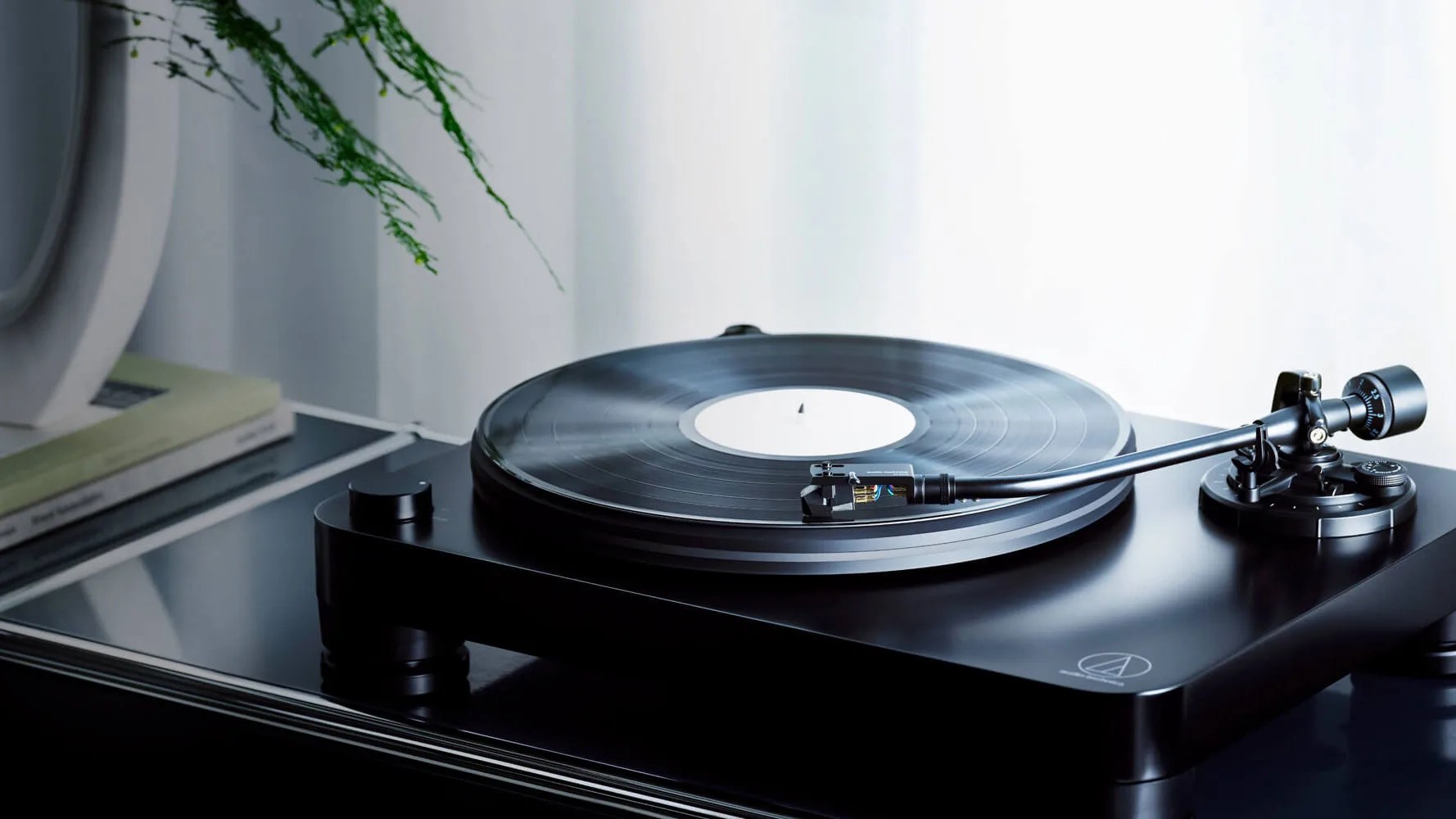 Black Audio-Technica turntable with a vinyl record on a reflective black surface near books and a plant.