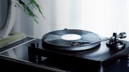 Black Audio-Technica turntable with a vinyl record on a reflective black surface near books and a plant.