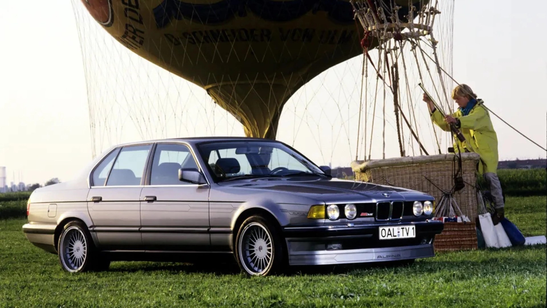 Silver BMW Alpina sedan parked on grass near a hot air balloon basket with a person in a yellow jacket.