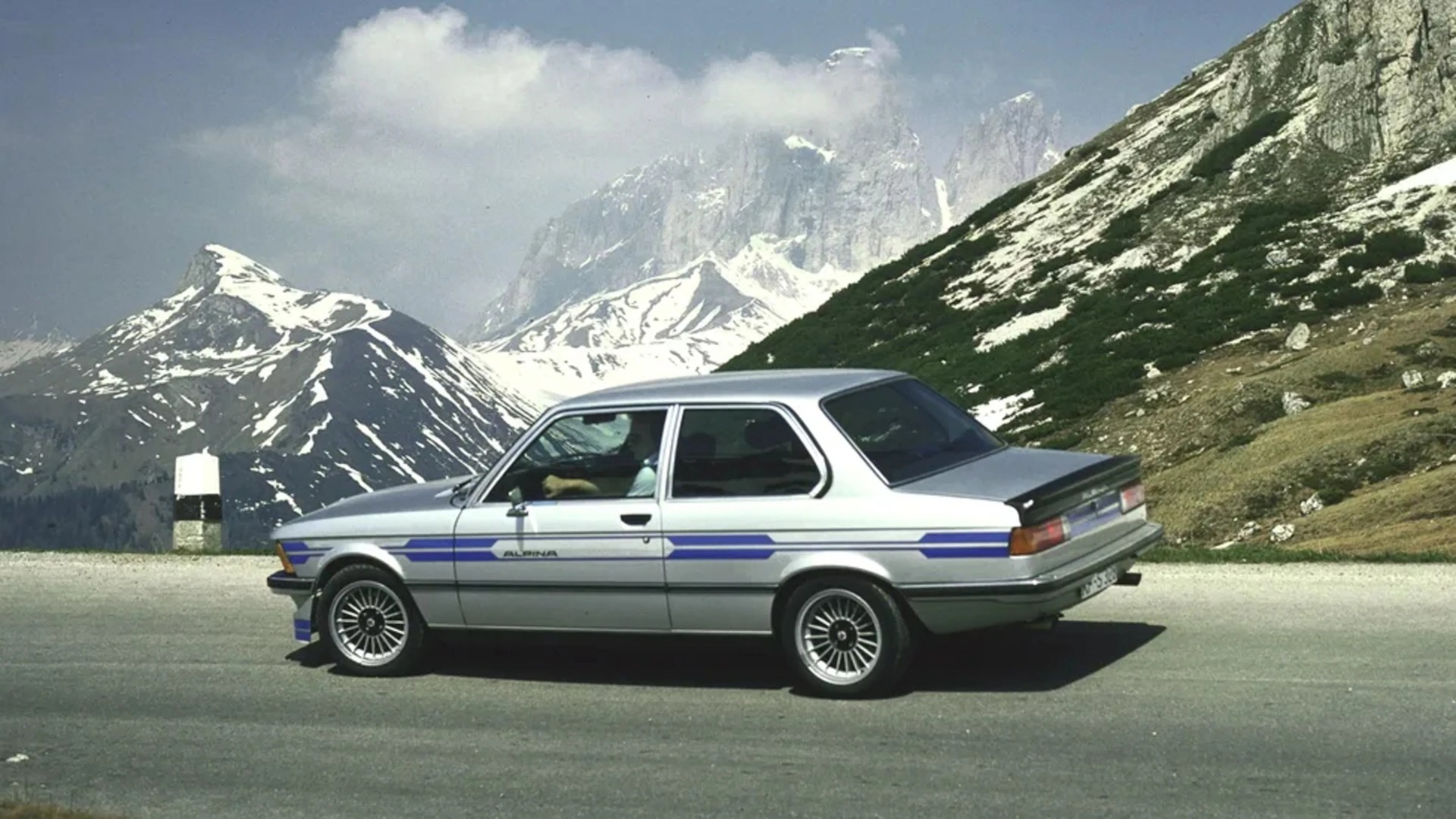 Silver two-door Alpina car with blue stripes parked on a mountain road with snow-capped peaks in the background.