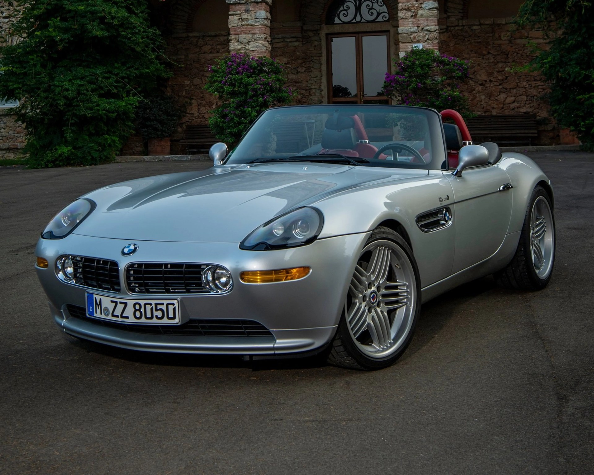 Silver BMW Z8 convertible with red interior parked on a driveway in front of a stone building.