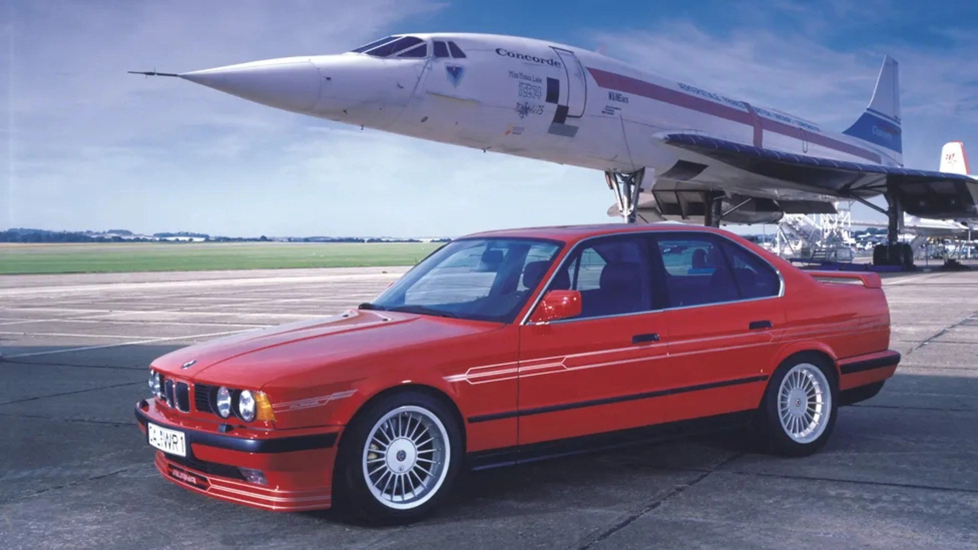 Red BMW Alpina sedan parked on an airport tarmac with a Concorde airplane in the background.