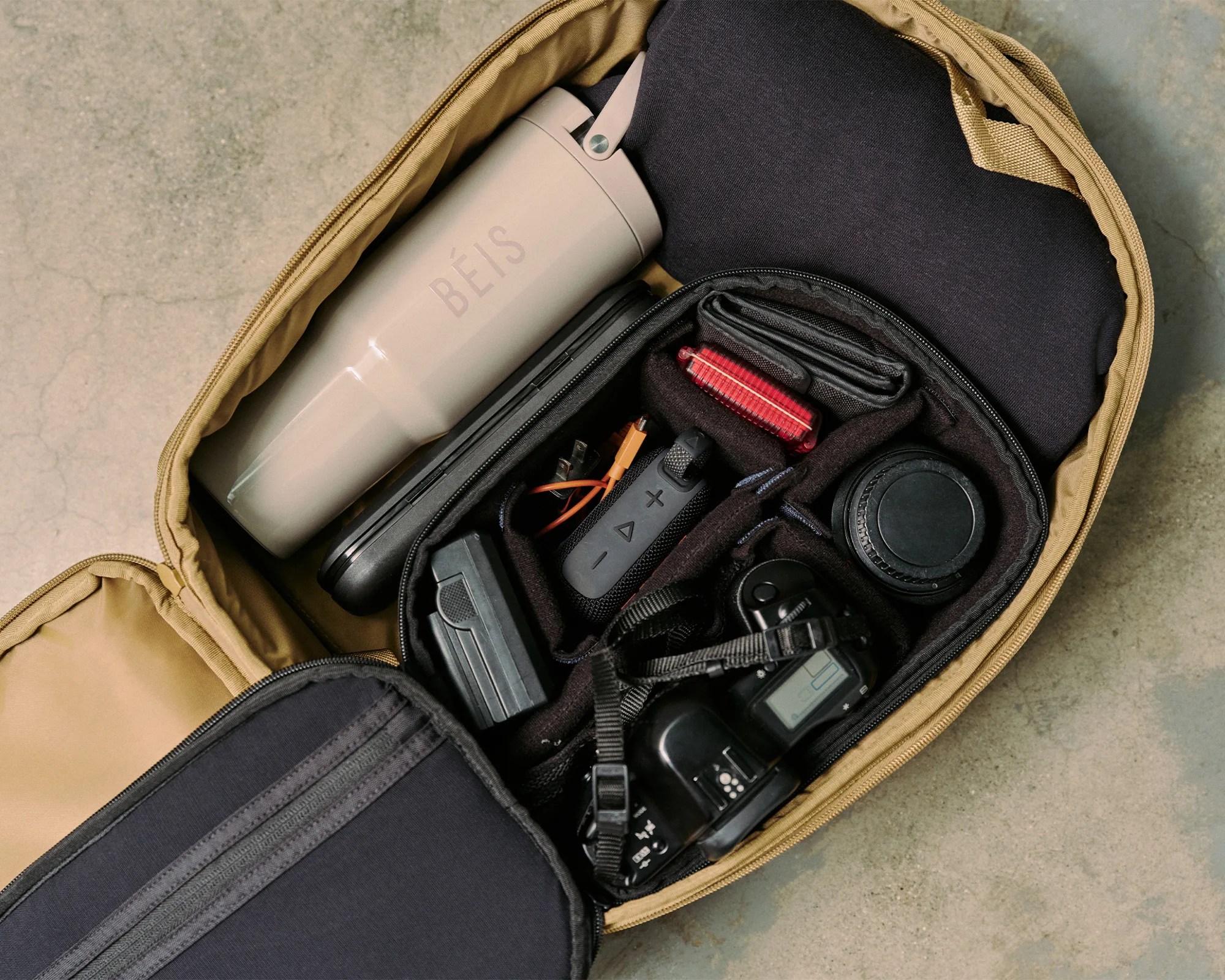 Tan camera bag with padded compartments holding a black camera, lens, cables, and a beige BÉIS water bottle.