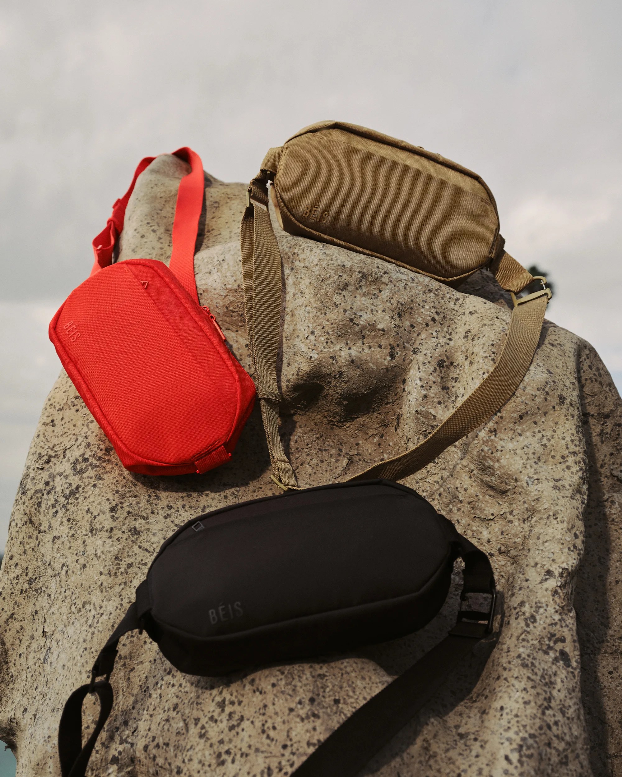 Three rectangular crossbody bags in red, tan, and black with adjustable straps on a large speckled rock.