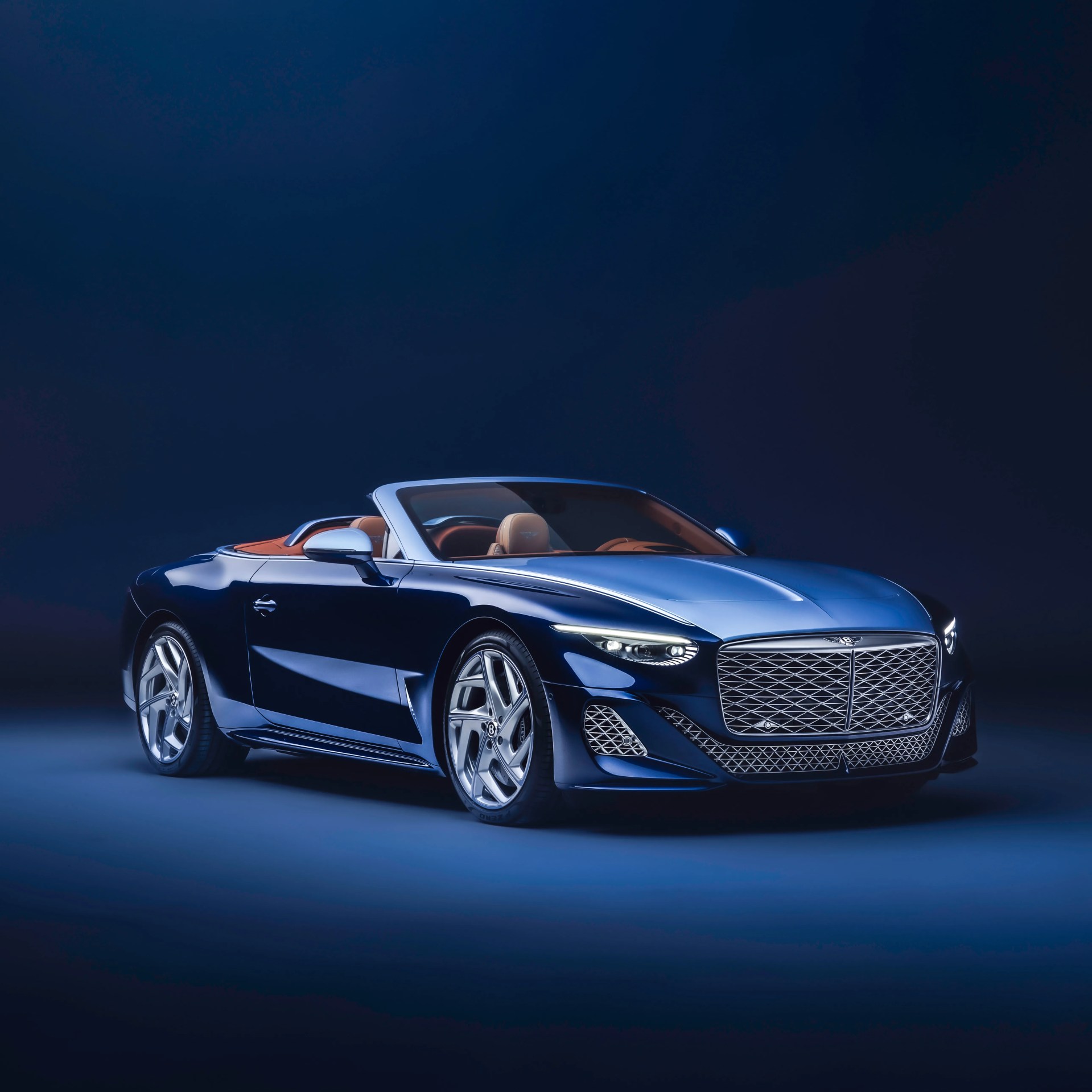 Dark blue Bentley convertible with tan leather interior and large mesh grille on a dark background.